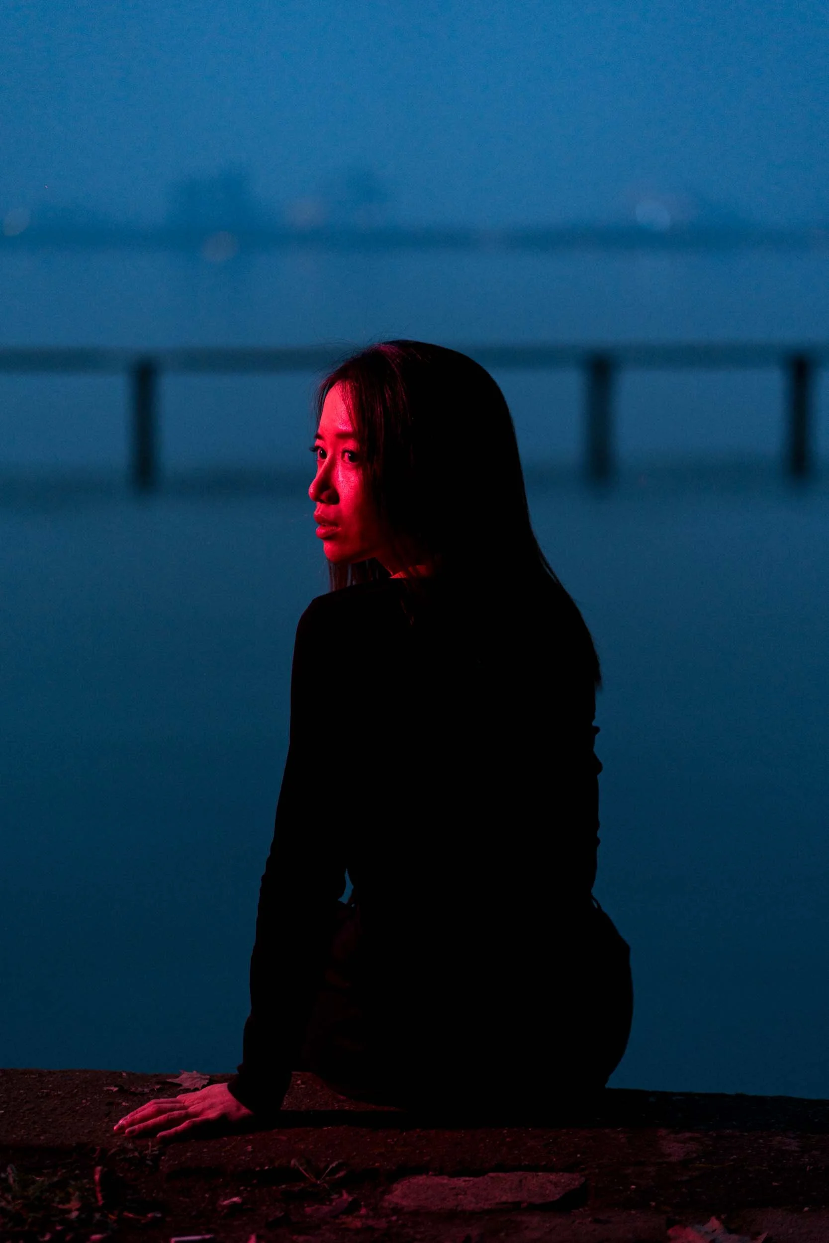 A woman with long dark hair kneeling on the ground at dusk or night, illuminated by pinkish-red lighting, with a dark blue sky and a bridge in the background.