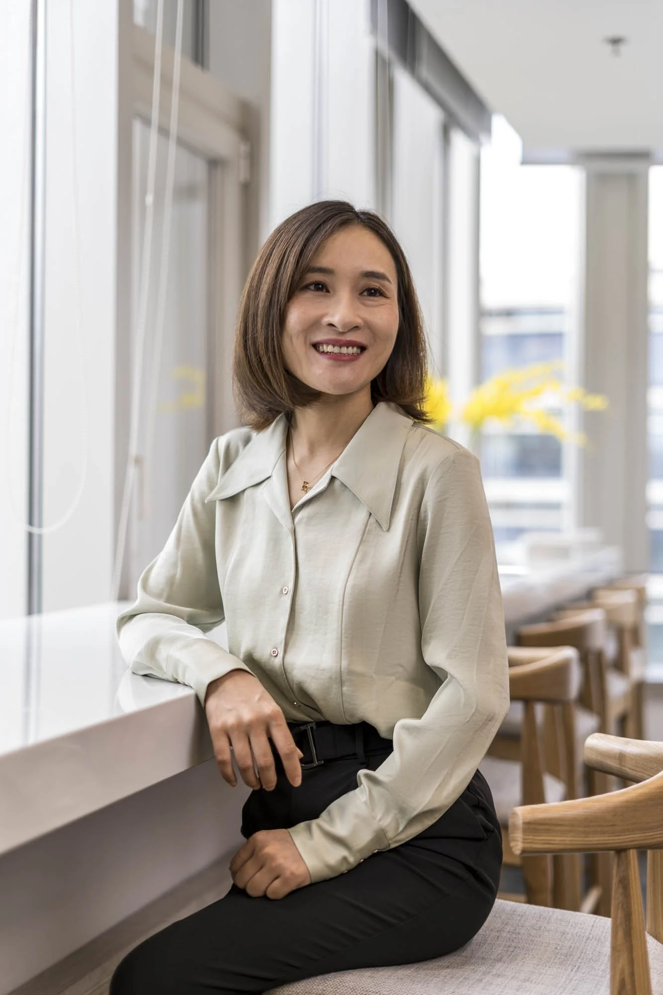 A woman with shoulder-length brown hair and a beige blouse sitting on a gray cushion by a white counter in a bright, modern setting with large windows and wooden chairs in the background.