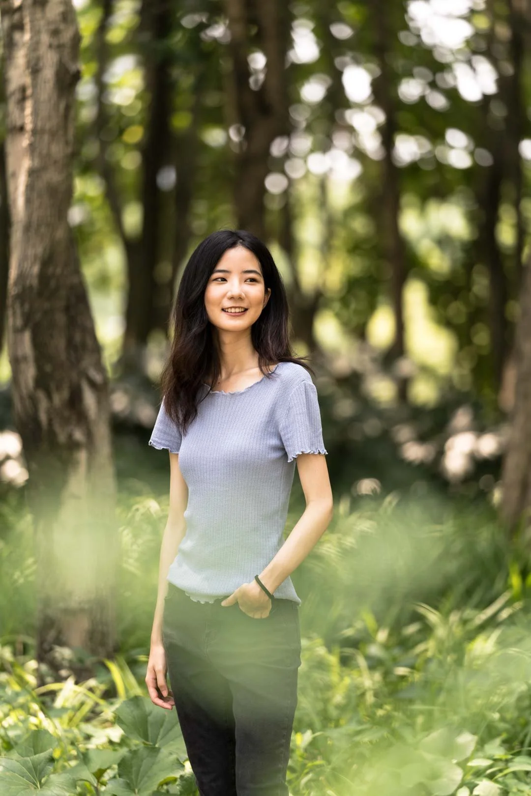 A woman with dark hair wearing a light gray top and black pants, standing outdoors among trees and greenery, smiling and looking to her right.