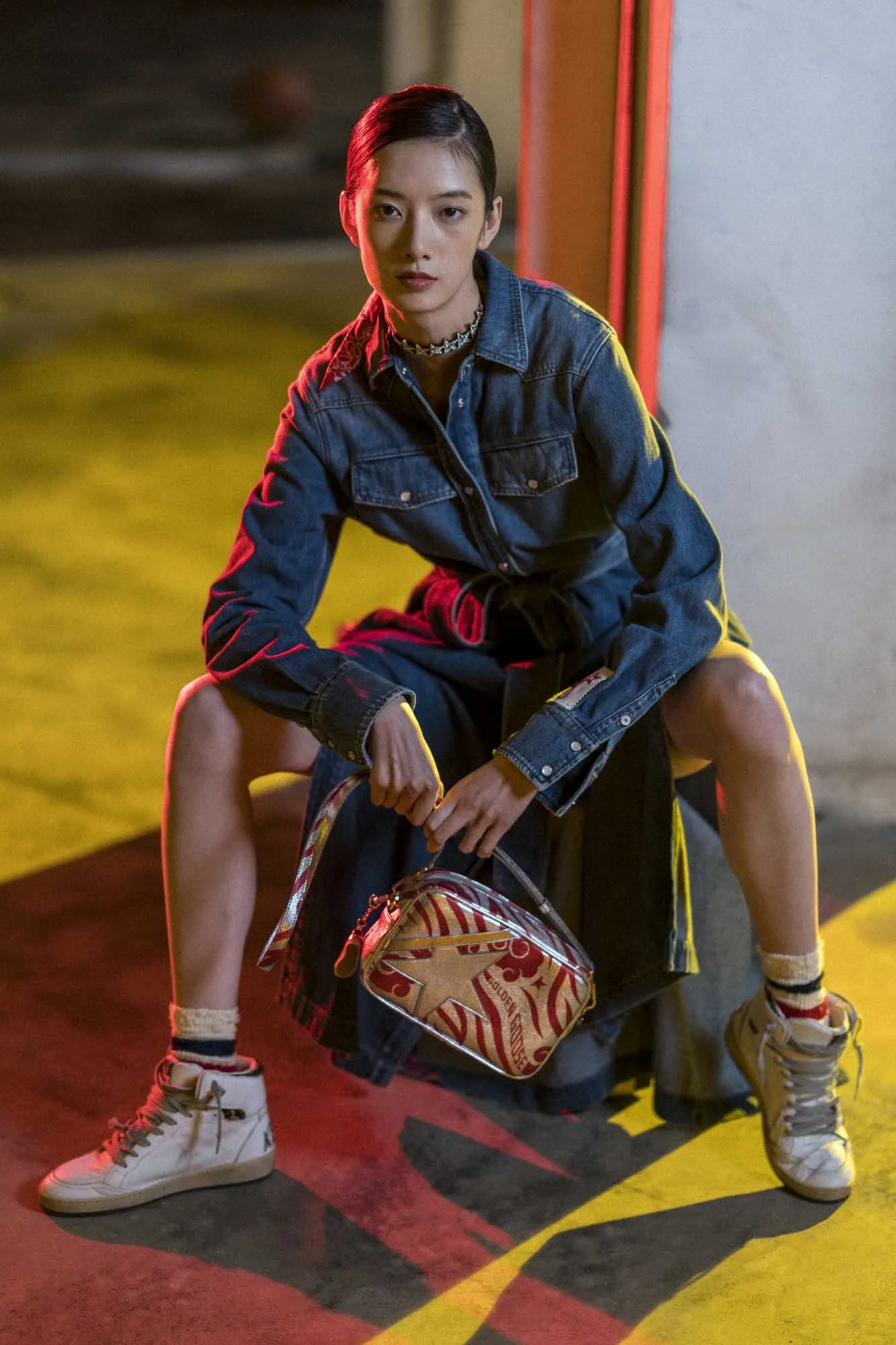 A young woman with short, dark hair and a choker necklace, wearing a denim jacket, is kneeling on a person with multiple legs on the ground in a dimly lit area. The scene has red and yellow lighting, and she is holding a bag with a red and beige graphic design.