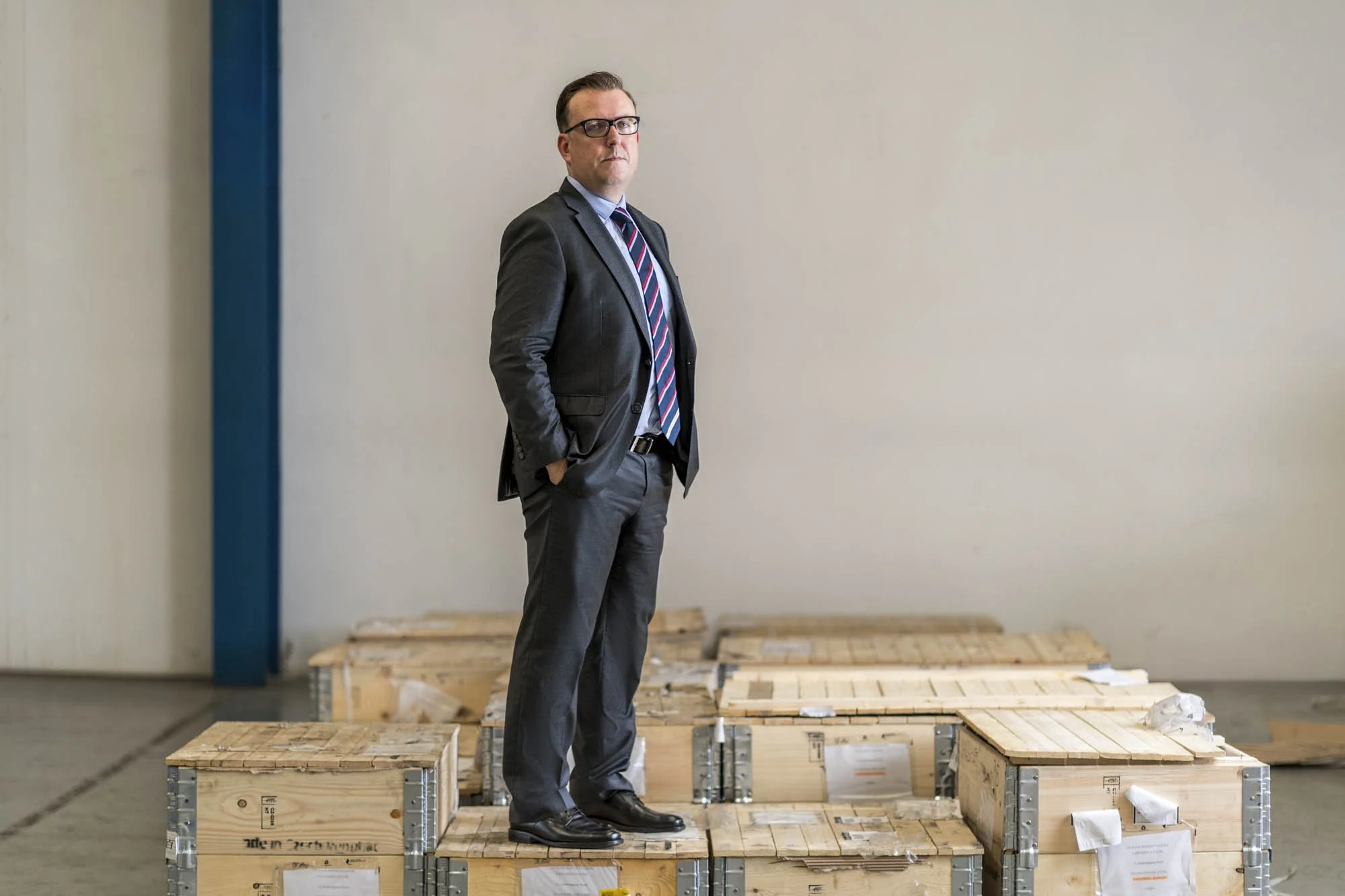 A man in a business suit standing among wooden crates, with a neutral wall and blue pillar in the background.