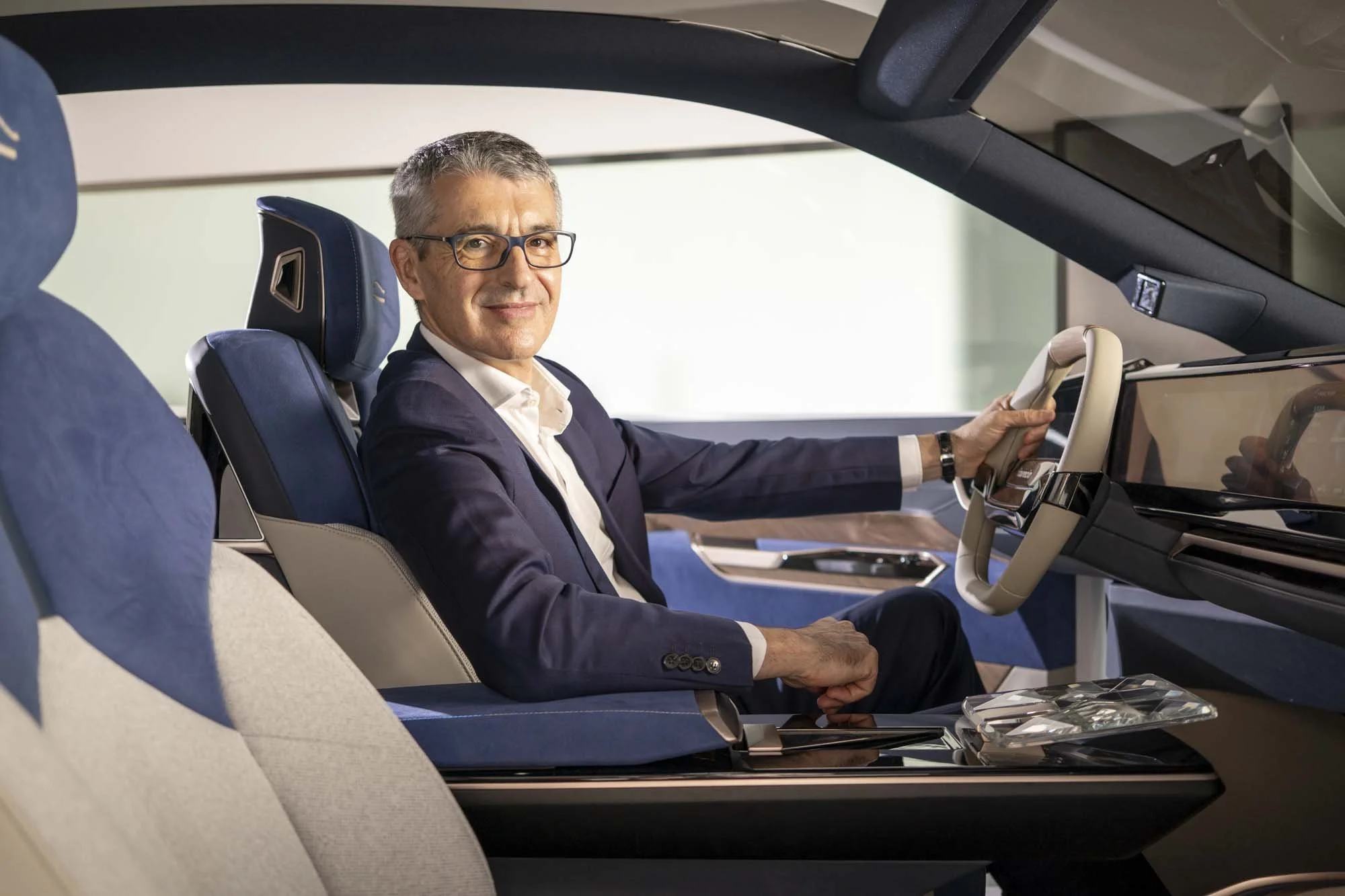 A man in a suit with glasses sitting in the driver's seat of a modern vehicle, smiling, with hands on the steering wheel.
