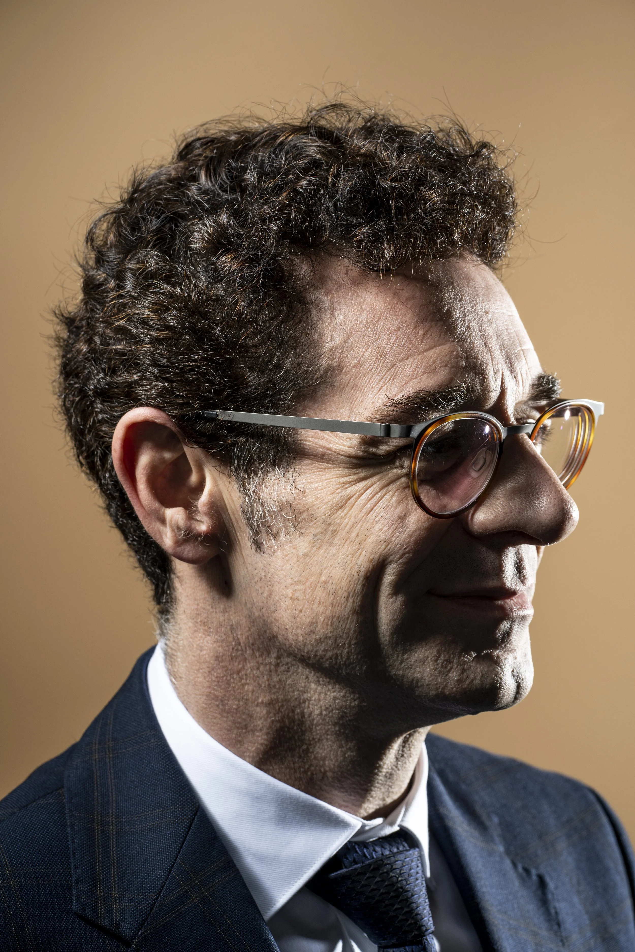 Close-up of a mature man with curly dark hair, wearing glasses, a dark checkered suit, white shirt, and dark tie, against a plain beige background.