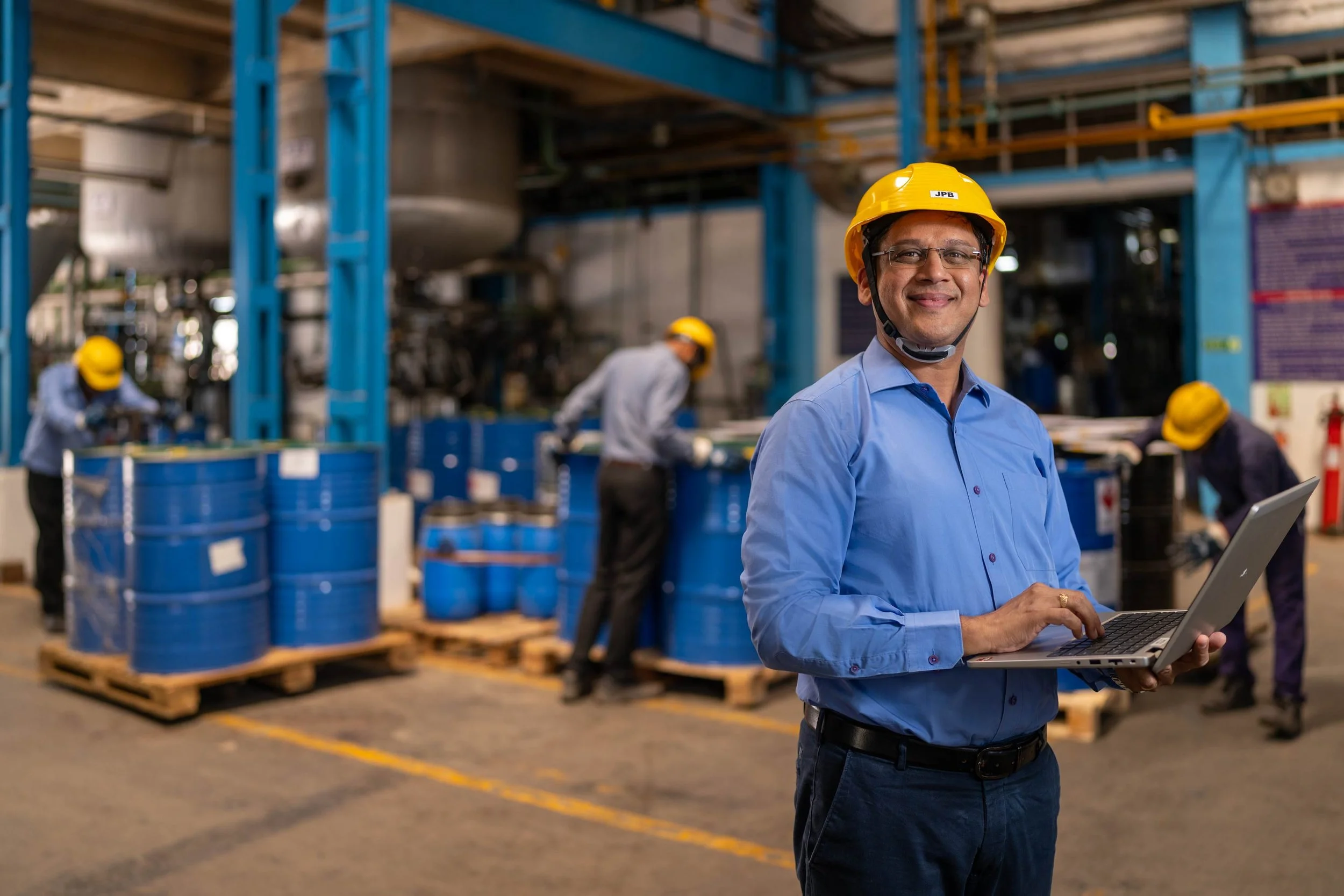 A man wearing glasses, a yellow safety helmet labeled 'JPB', and a blue dress shirt, holds a laptop while standing in a warehouse with industrial shelving and workers in the background.