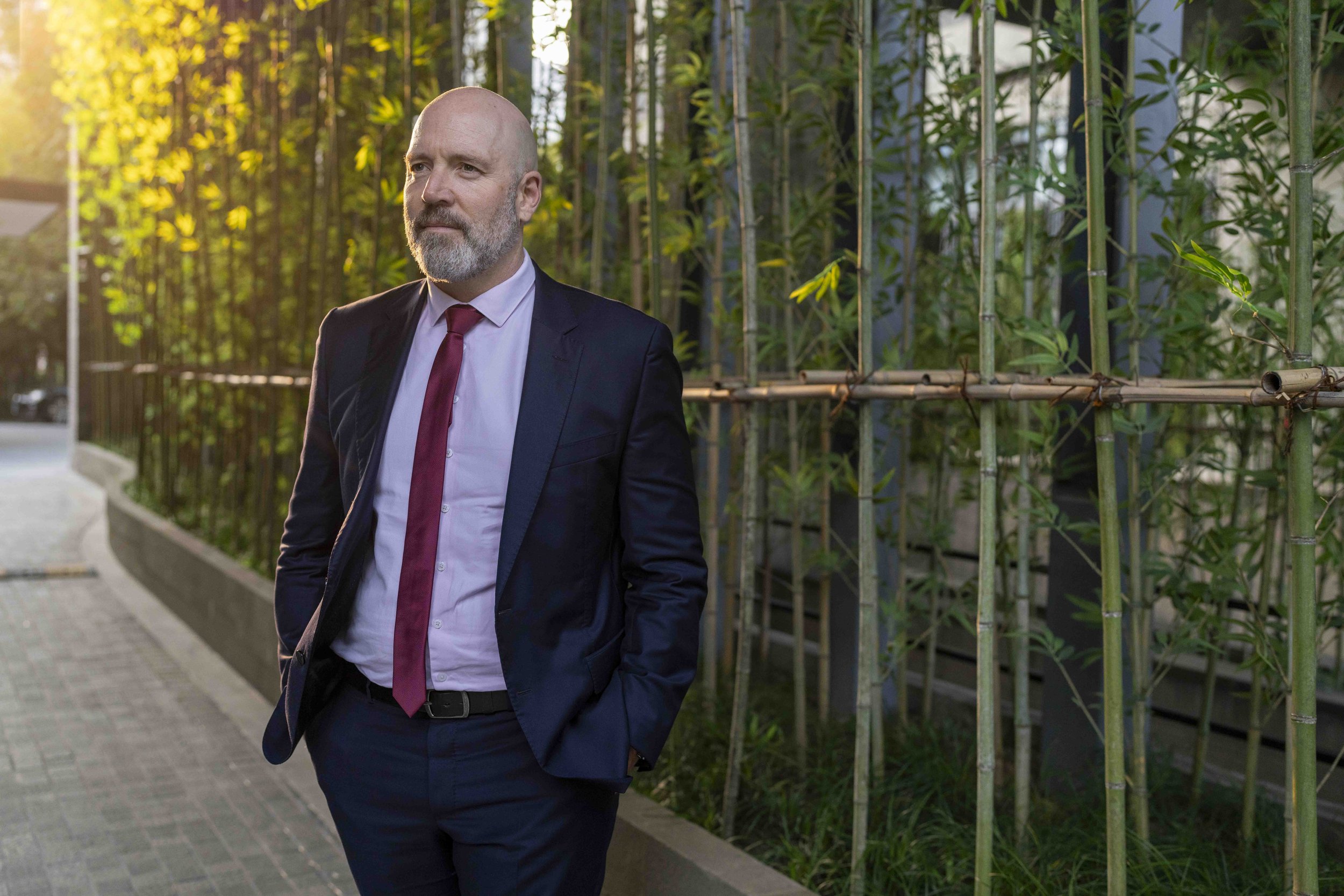 A bald man with a gray beard wearing a dark suit, white shirt, and red tie standing outdoors in front of a green bamboo fence during sunset.