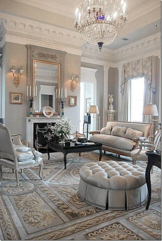 For modern tastes, there is not much desire for clutter, but a sense of opulence is still created using similar ideas to those in Victorian times. Deep buttoned upholstery, gilding, a thick rug and glass chandelier give a sense of luxury. The multi-layered widow treatment, mounted urn and emphasis on the room's ceiling height brings grandeur. The lighting is soft and the reflective surface of the coffee table, as well as the brass wall lights, candlesticks and mirror will ensure this room sparkles as evening sets in. The limited colour palette is lifted through this clever use of accessories and texture.