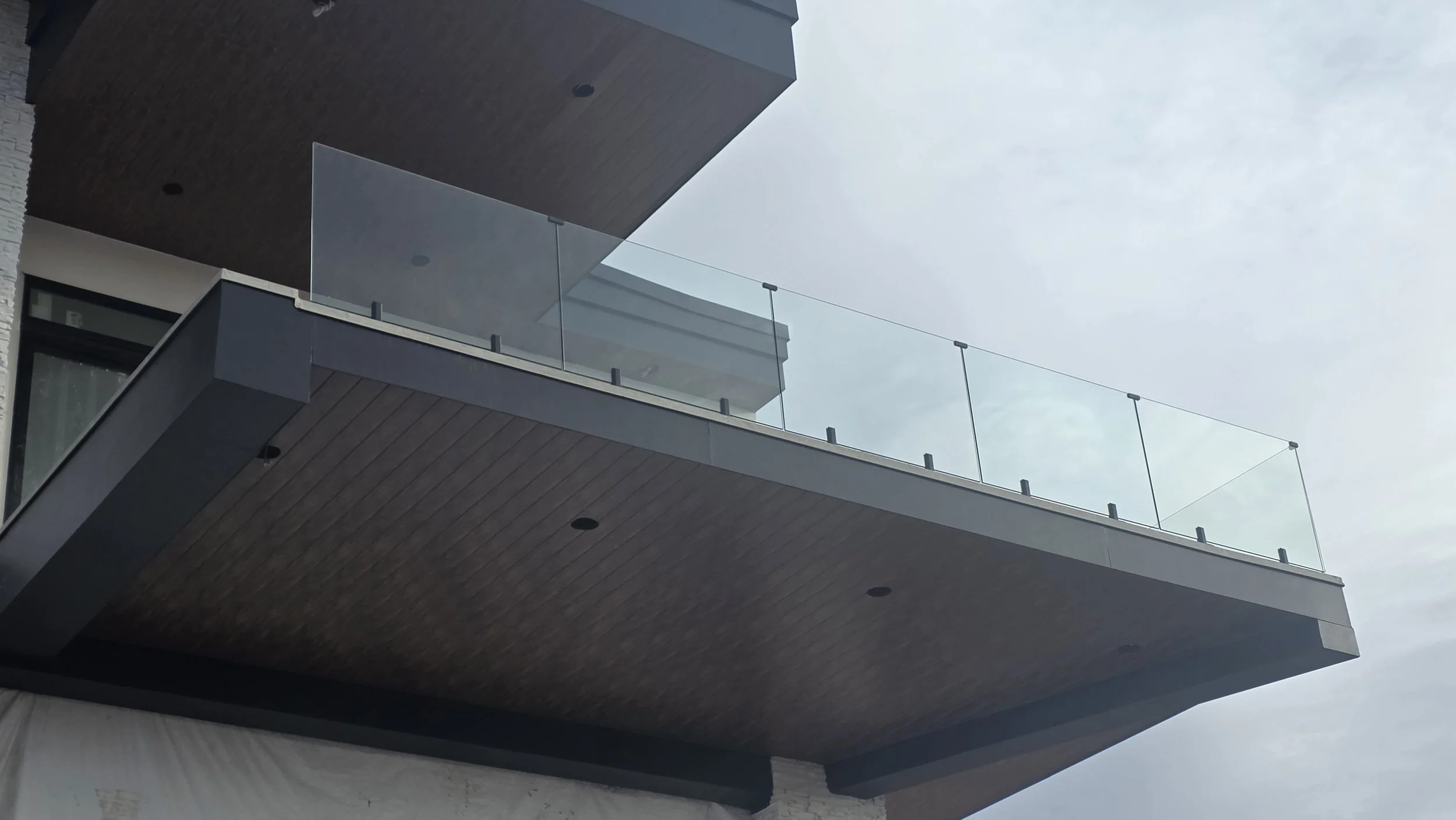 View of a modern building's balcony with glass railing and wooden ceiling, against a cloudy sky.