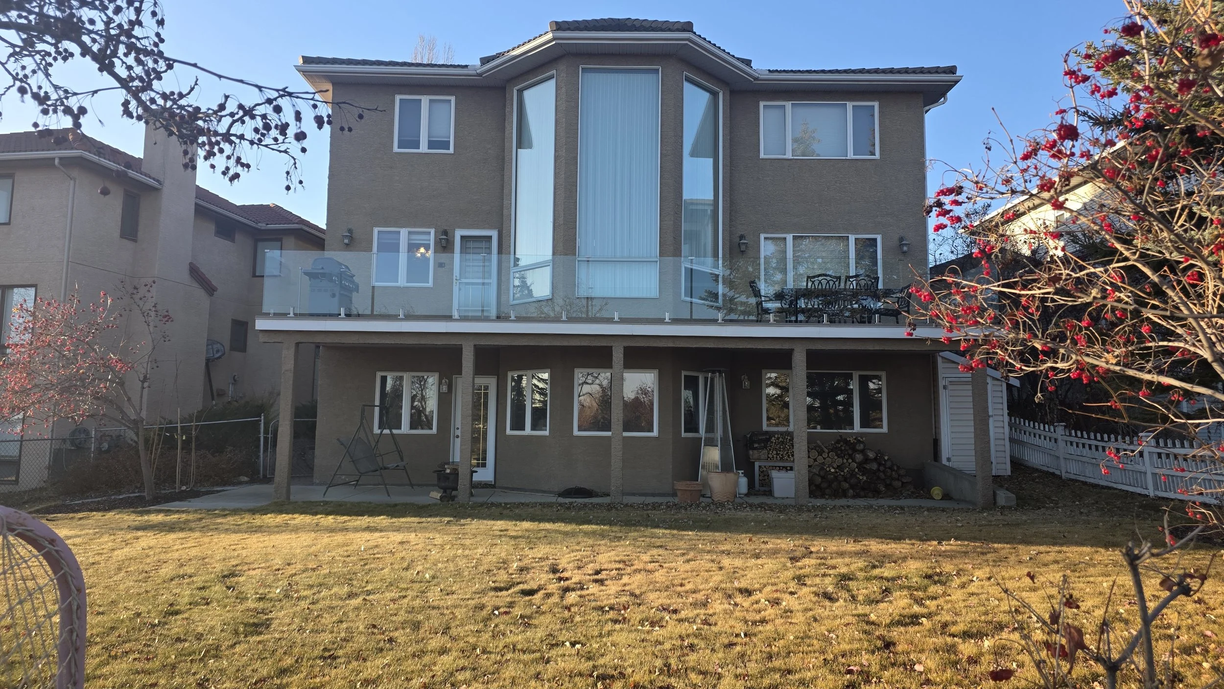 A multi-story house with beige exterior walls, multiple windows, and balconies featuring glass railings, situated on a grassy yard with trees and shrubs.