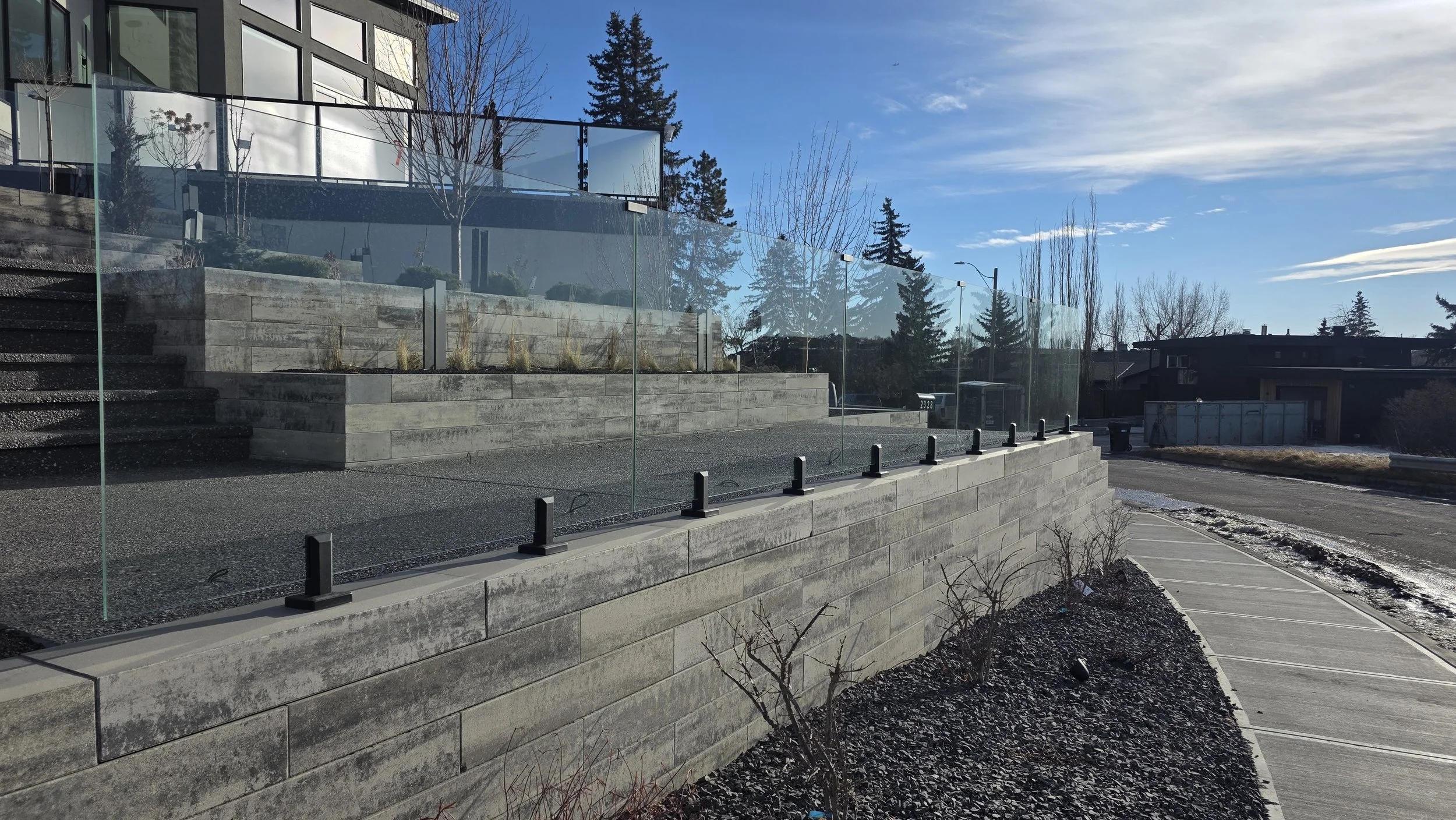 A modern residential exterior featuring a glass fence on a multi-level concrete patio with steps, a driveway, leafless bushes, and a clear blue sky.
