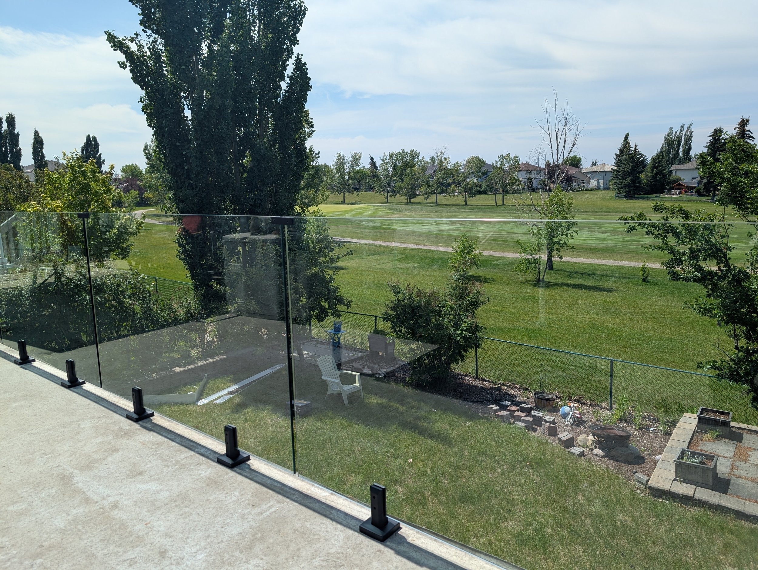 View of a backyard with a glass railing, surrounded by trees, grass, and houses in the distance on a partly cloudy day.