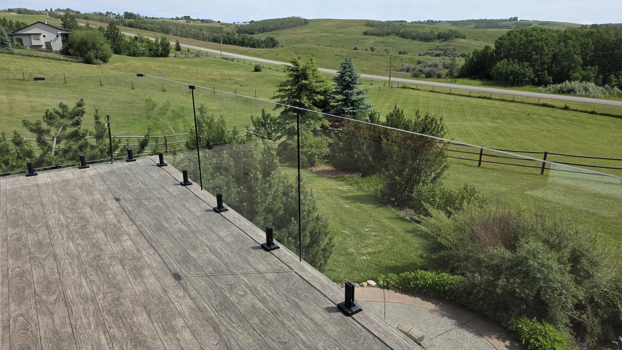 View from a wooden deck with a glass railing overlooking a green backyard with trees and a wide open field in the distance.