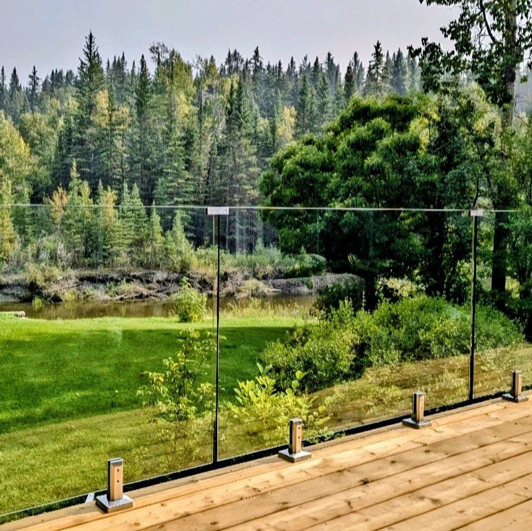 Glass railing on a wooden deck overlooking a grassy yard, pond, dense trees, and forested landscape.