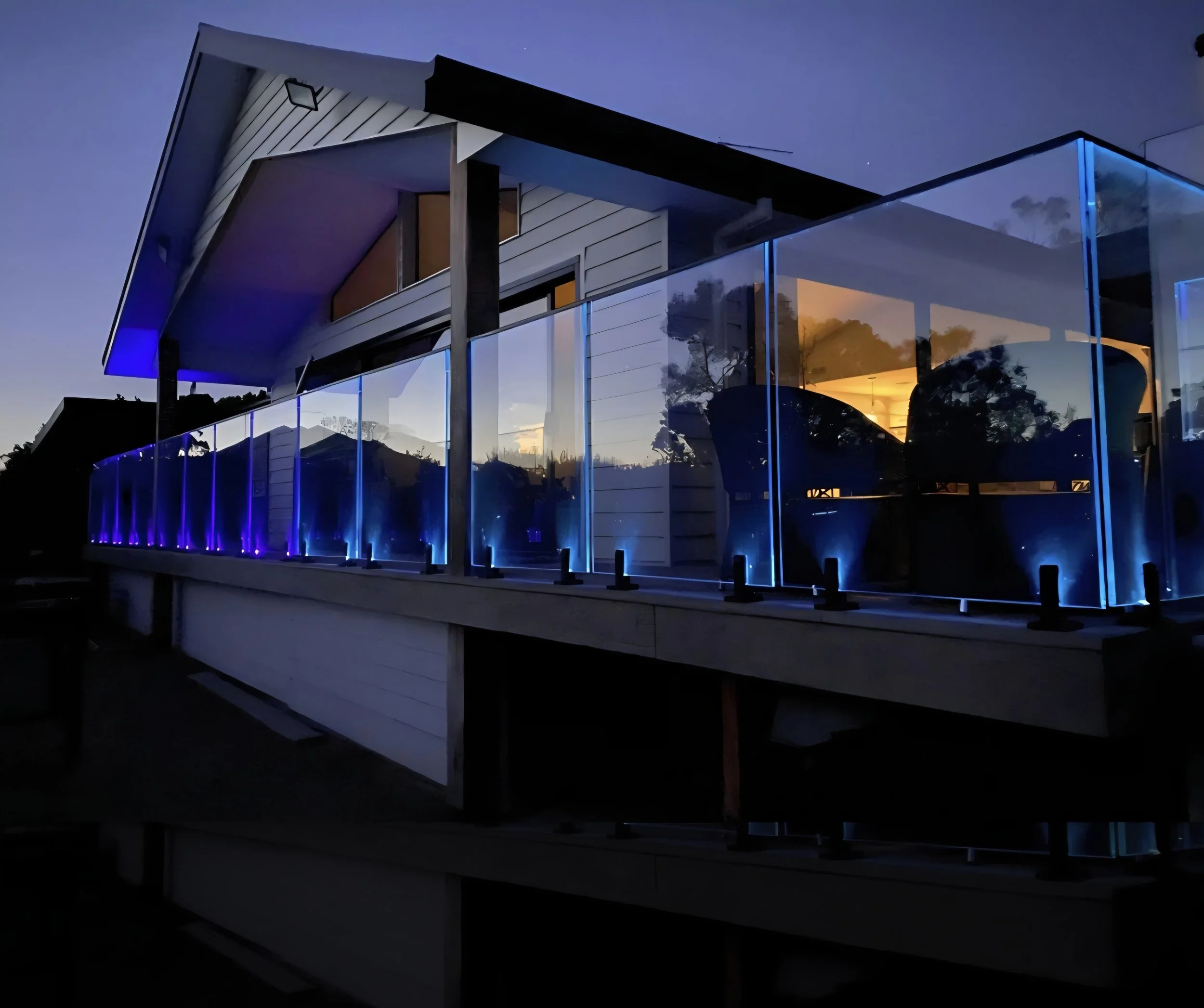Modern house with glass balcony railing illuminated by blue and yellow lights at dusk.