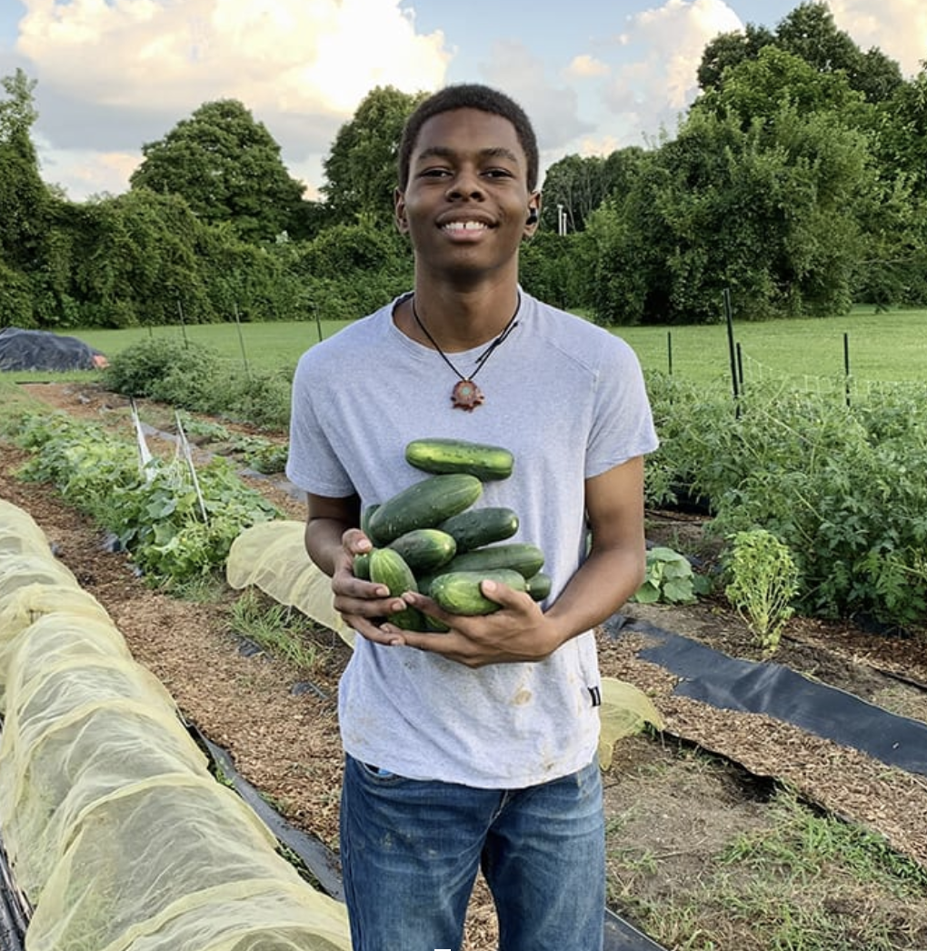 A Young Eco-Hero Award Winner smiling and holding cucumbers in a garden with plants and trees in the background.