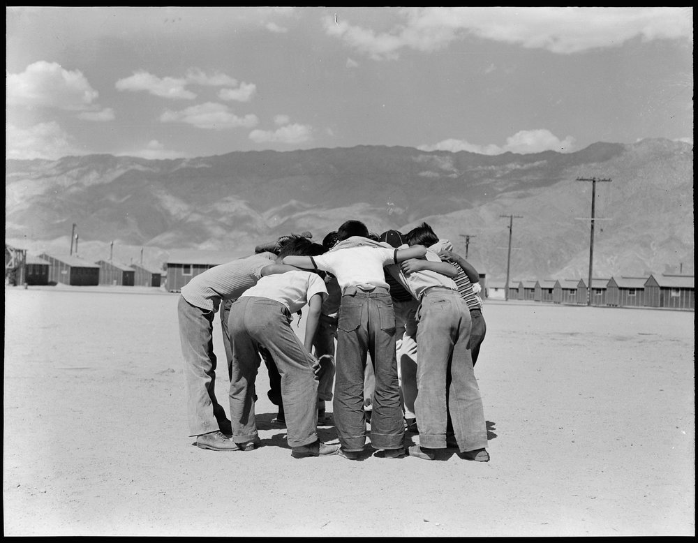  Manzanar Relocation Center, Manzanar, California. Baseball players in a huddle. This game is very popular with 80 teams having been formed to date. Most of the playing is done in the wide firebreak between blocks of barracks. 