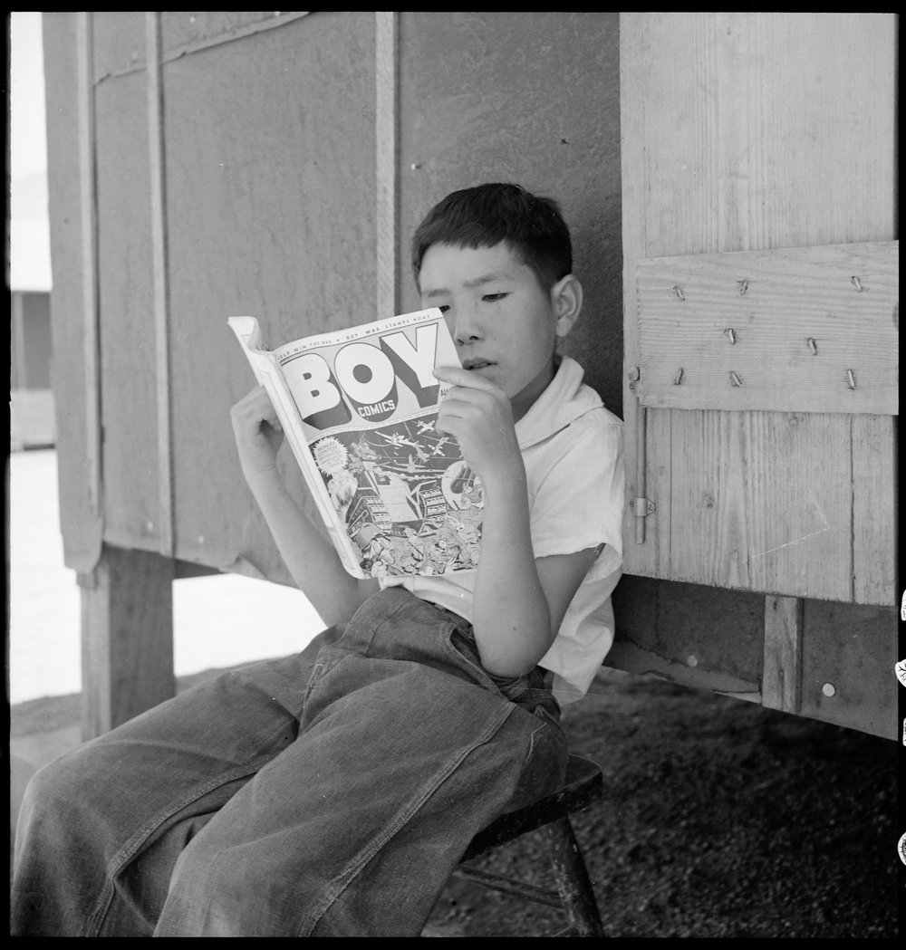  Manzanar Relocation Center, Manzanar, California. Evacuee boy at this War Relocation Authority center reading the Funnies. 