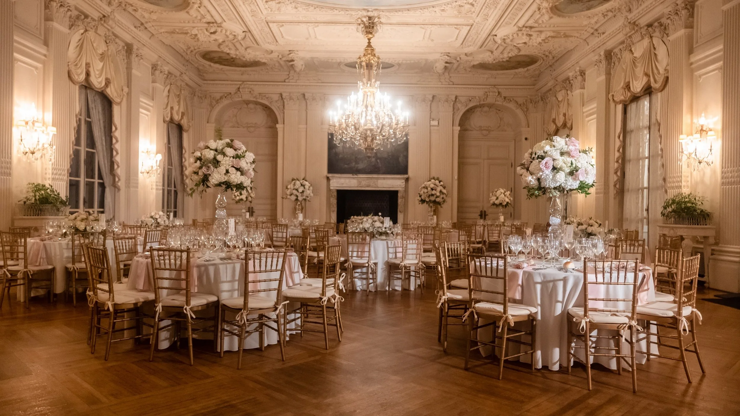 The ballroom at Rosecliff Mansion set for a wedding dinner, round tables laid in cream and blush beneath a lit crystal chandelier, tall florals on every table