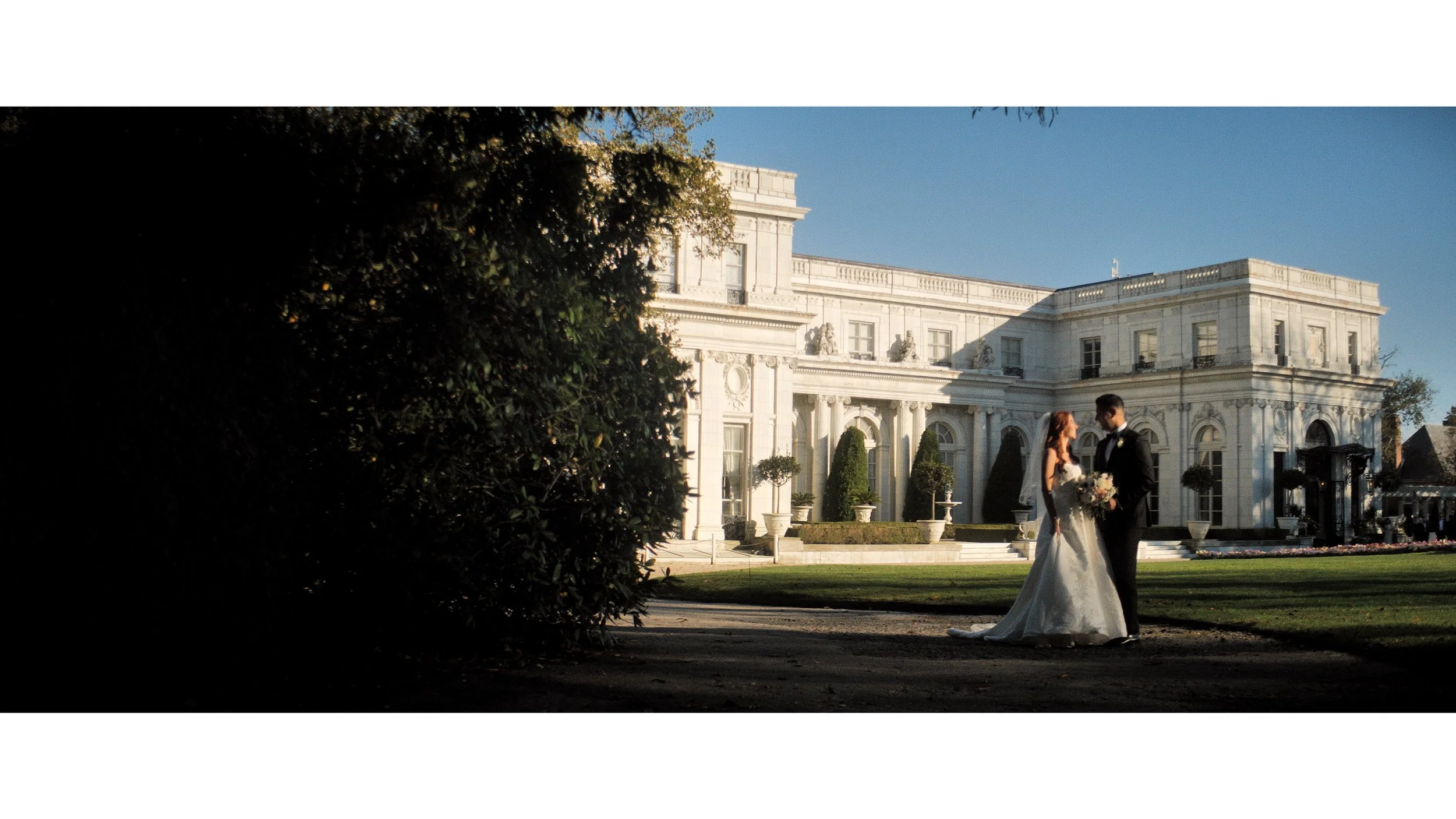 The arched garden front of Rosecliff Mansion in afternoon sun, a couple standing small on the lawn at the edge of the shade, Newport, Rhode Island