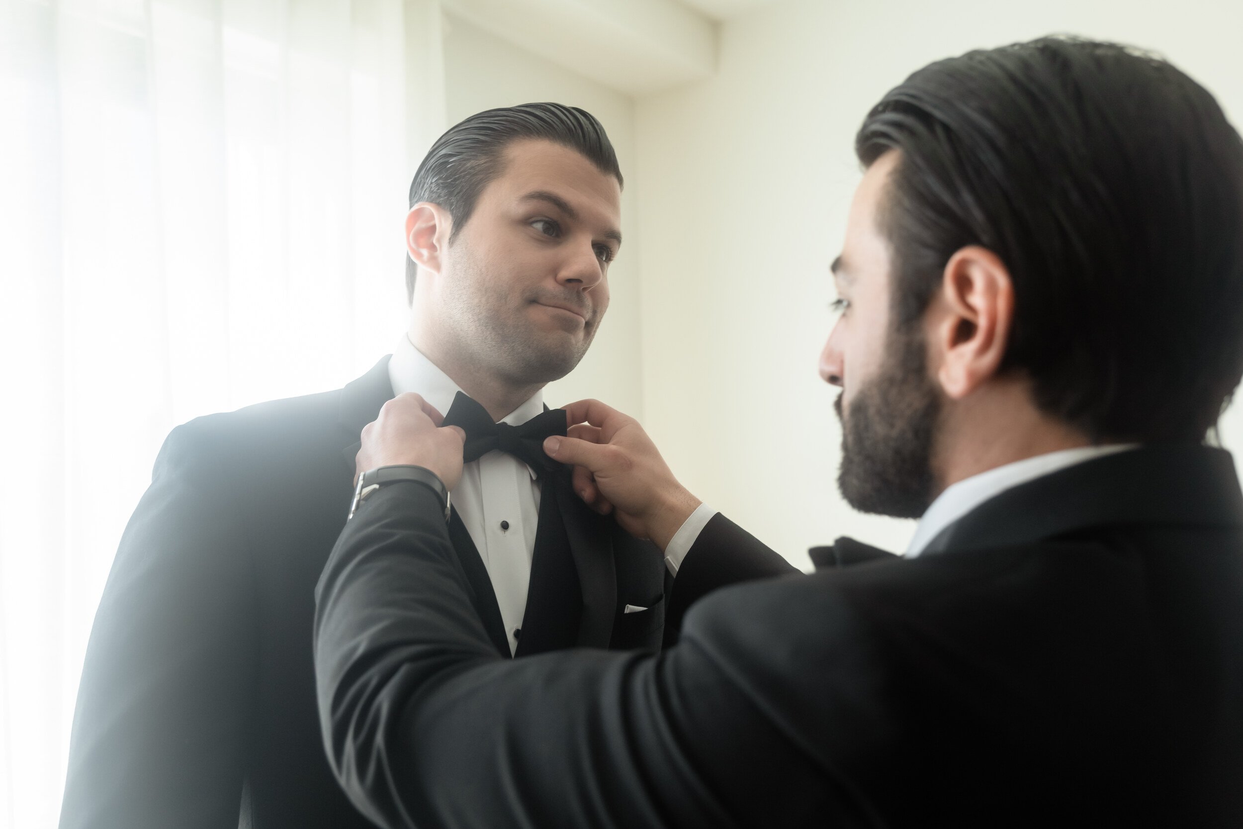 Groom in a tuxedo having his black bow tie straightened by another man in a tuxedo, window light, Wayfinder Hotel.