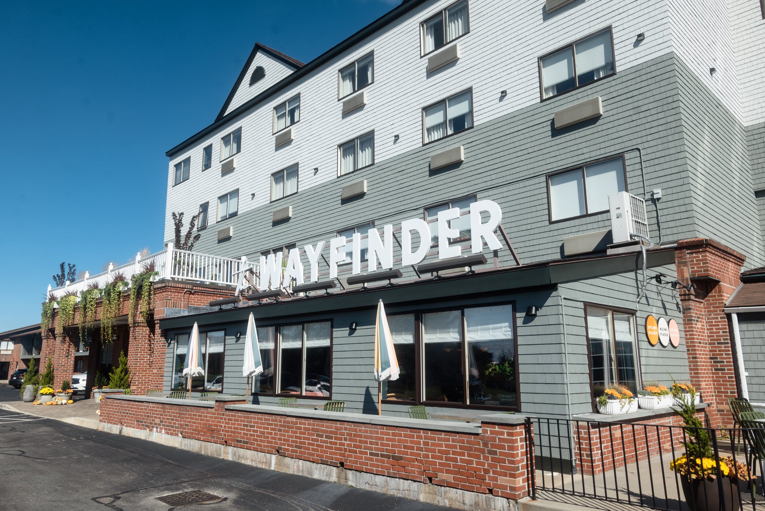 Exterior of the Wayfinder Hotel, its sign lit against a blue sky, autumn planters on the brick patio, no people.