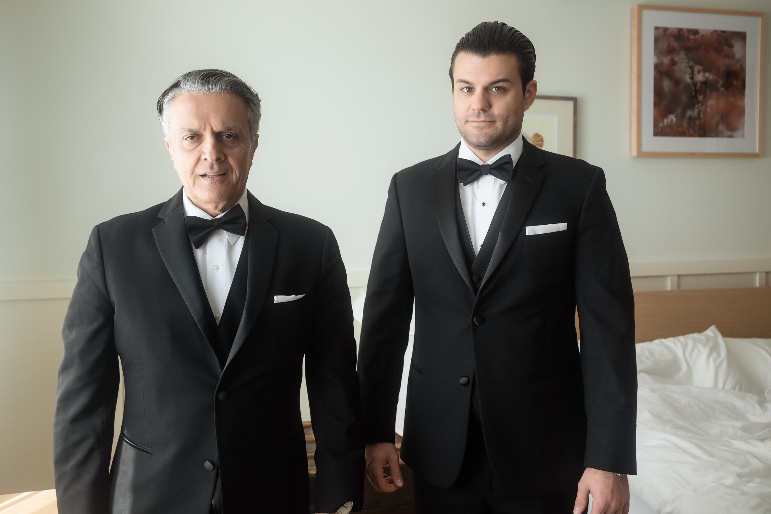 Groom in a tuxedo standing beside another man in a matching tuxedo in a hotel room, Wayfinder Hotel.