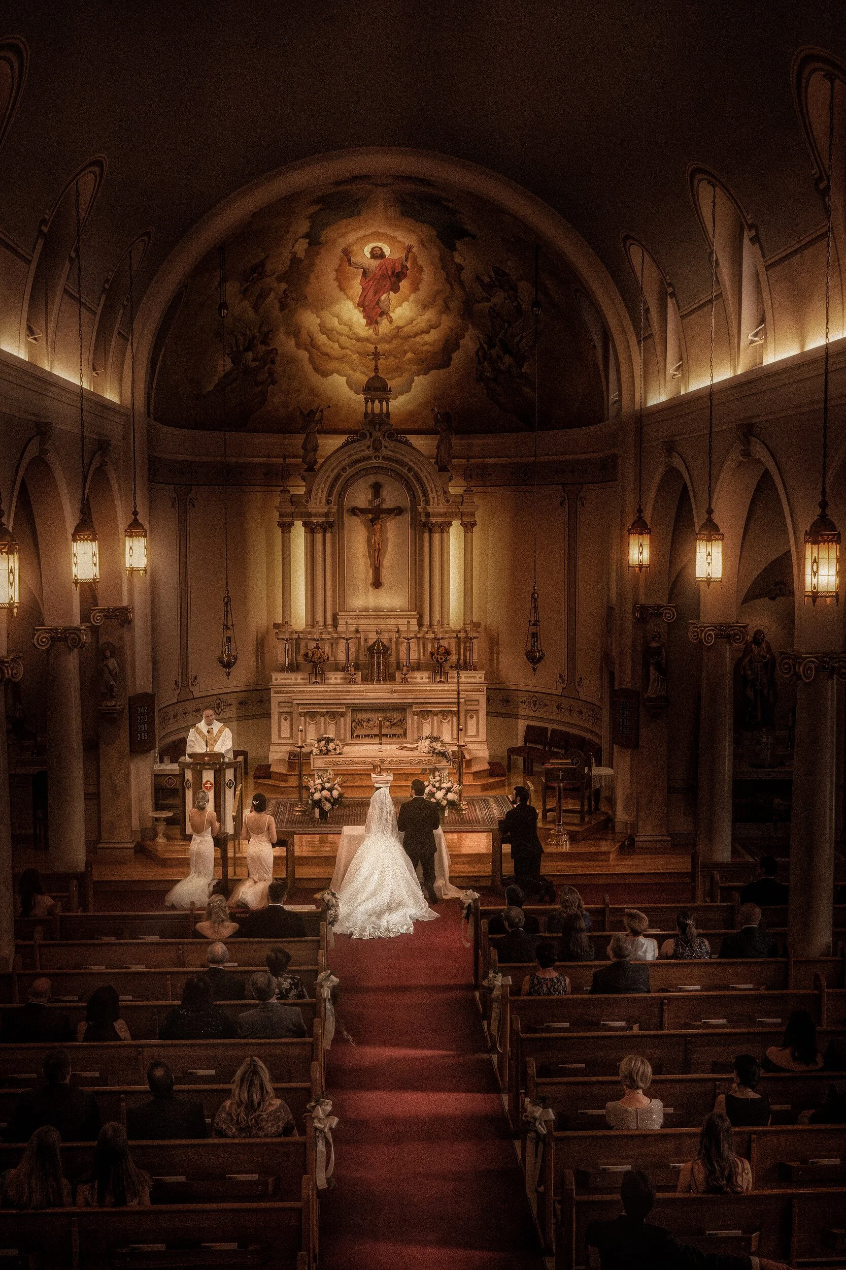 Wide interior view of the Church of Jesus Saviour during the ceremony, seen from the choir loft: the couple kneeling before the altar beneath a painted fresco of the Ascension, the full congregation in the pews below.