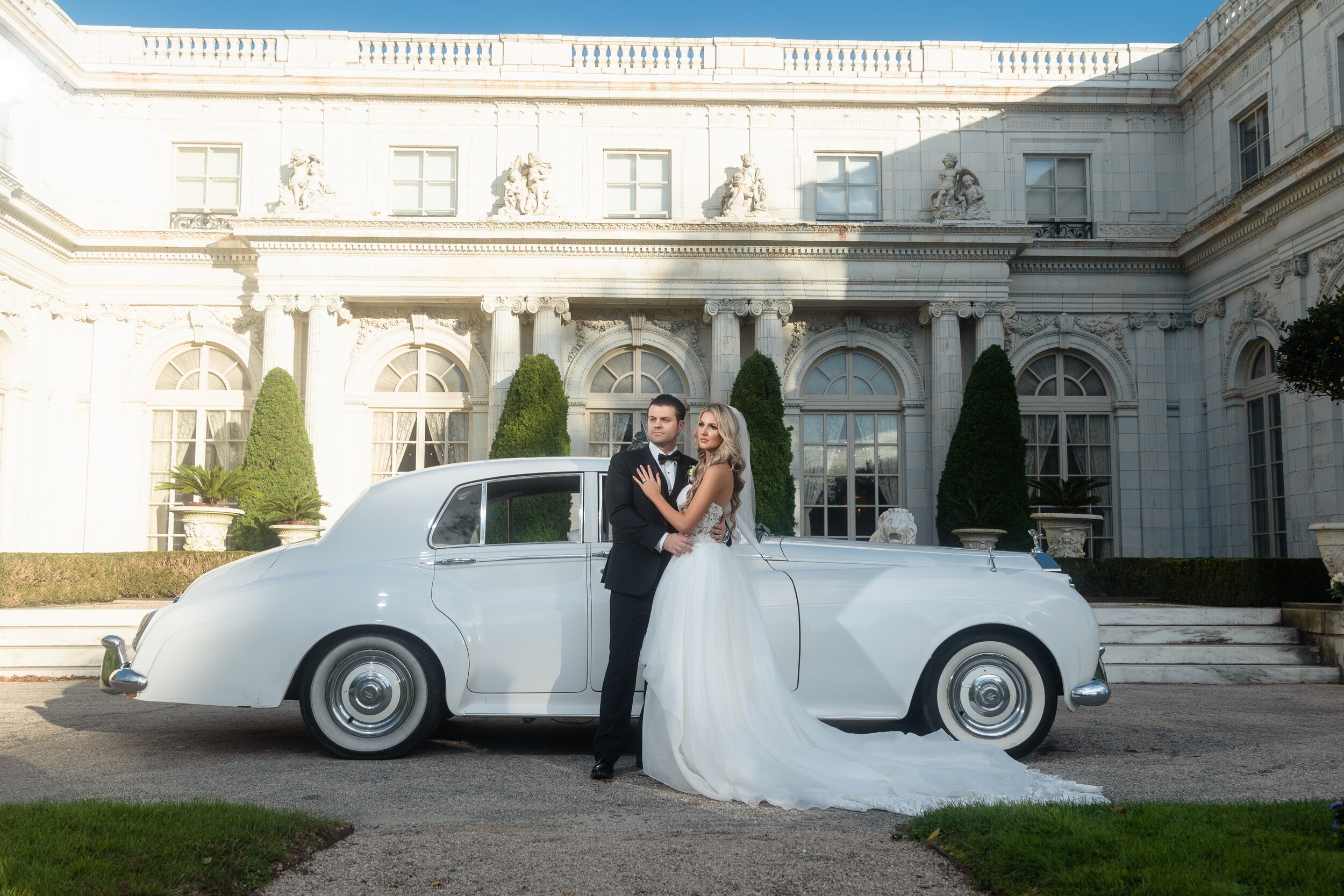 Gina and Anthony beside a vintage white Rolls-Royce in front of Rosecliff's marble facade, golden afternoon light, the mansion's columns and arched windows behind them.