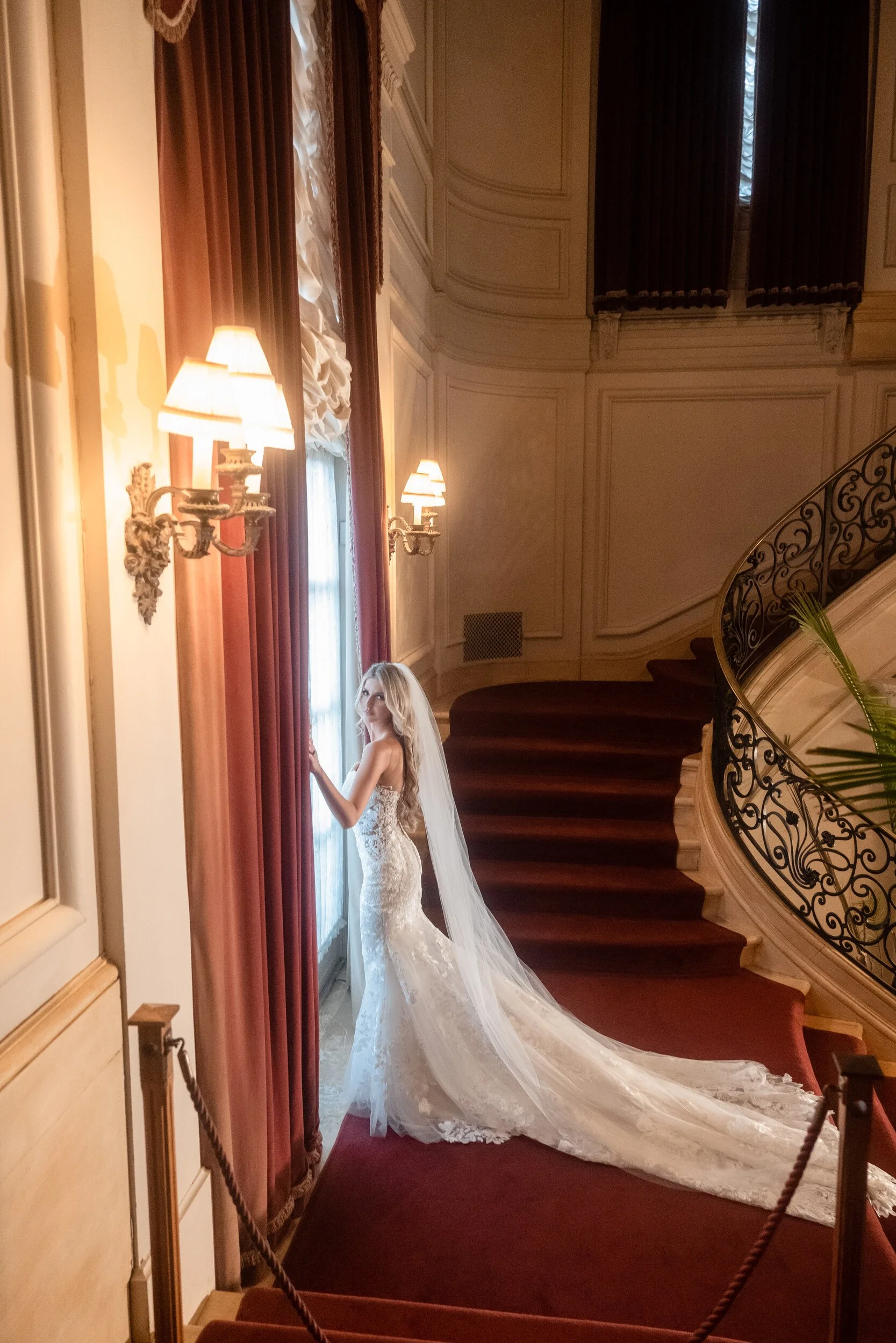 Gina pausing at a curtained window beside Rosecliff's curved grand staircase, red-carpeted steps and a wrought-iron railing behind her.