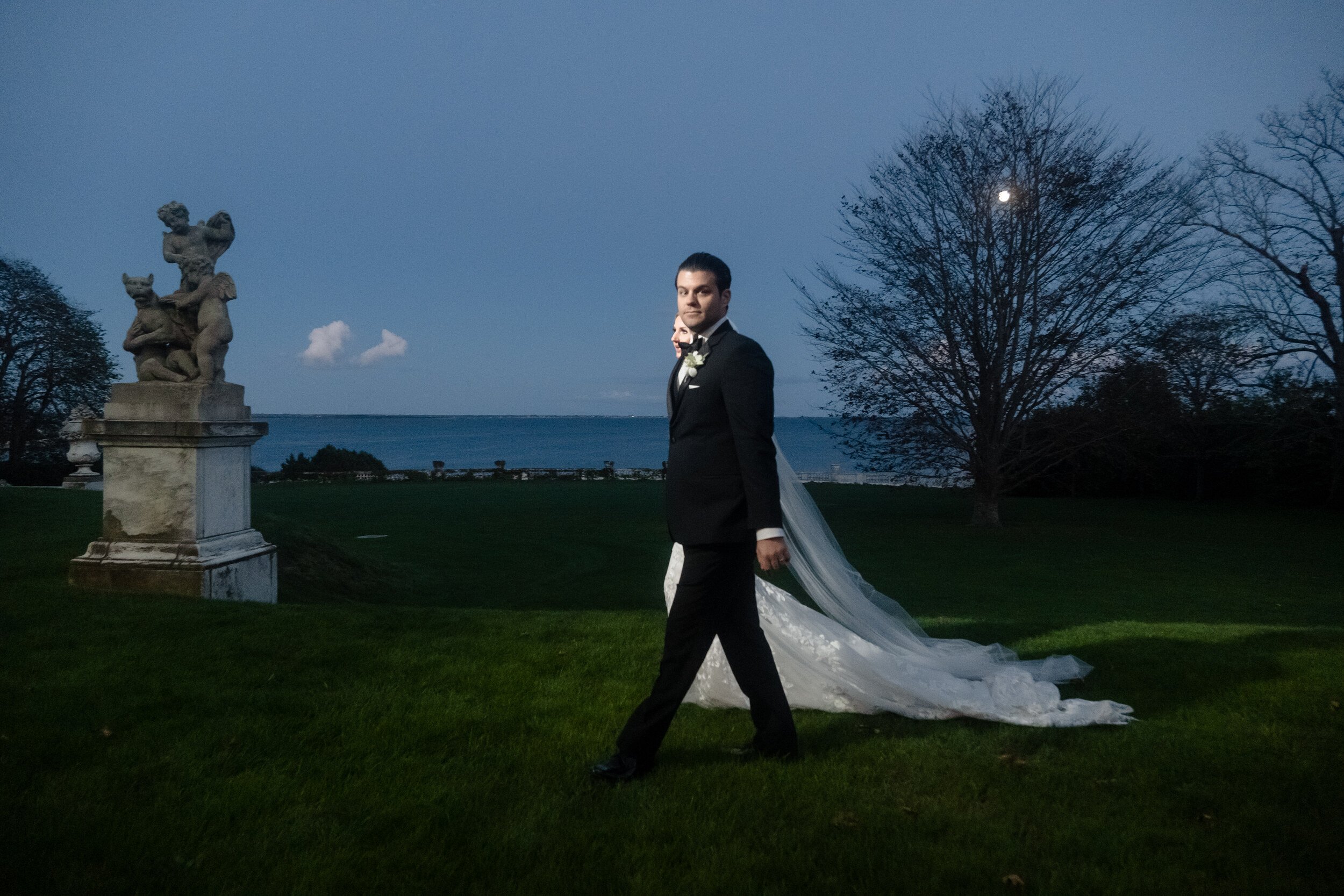 Groom walking across a dark lawn at Rosecliff with the bride close behind him, her face visible over his shoulder and her dress train trailing on the grass, the ocean and the moon behind them.