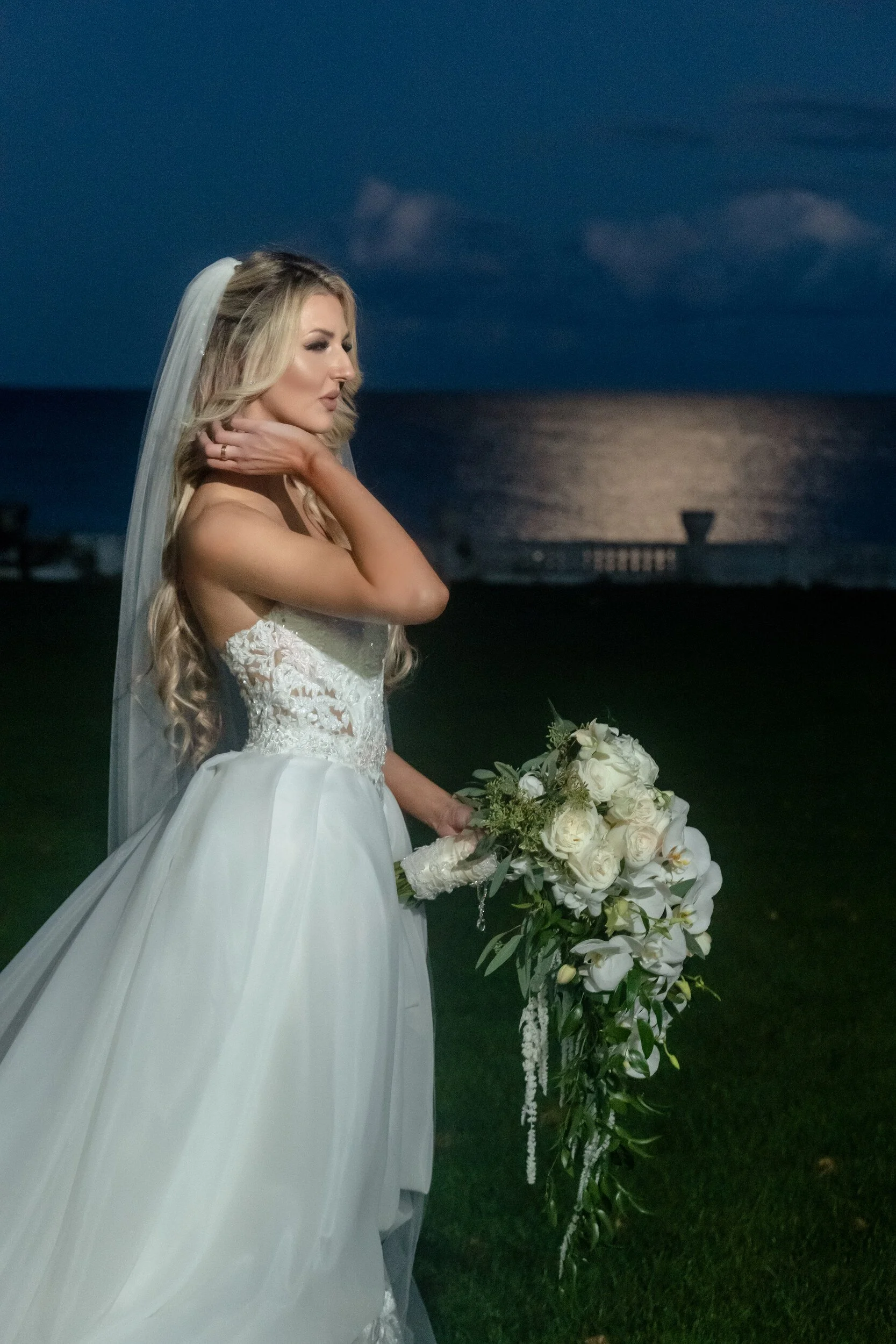 Gina in profile at dusk, hand at her collarbone, holding her white bouquet, the ocean behind her.