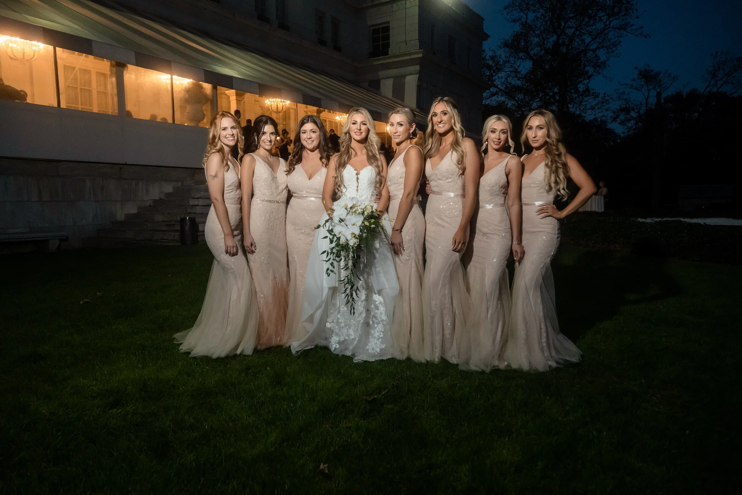 Bride and her bridesmaids standing in a line at night, the bride holding a white bouquet, Rosecliff lit up behind them.