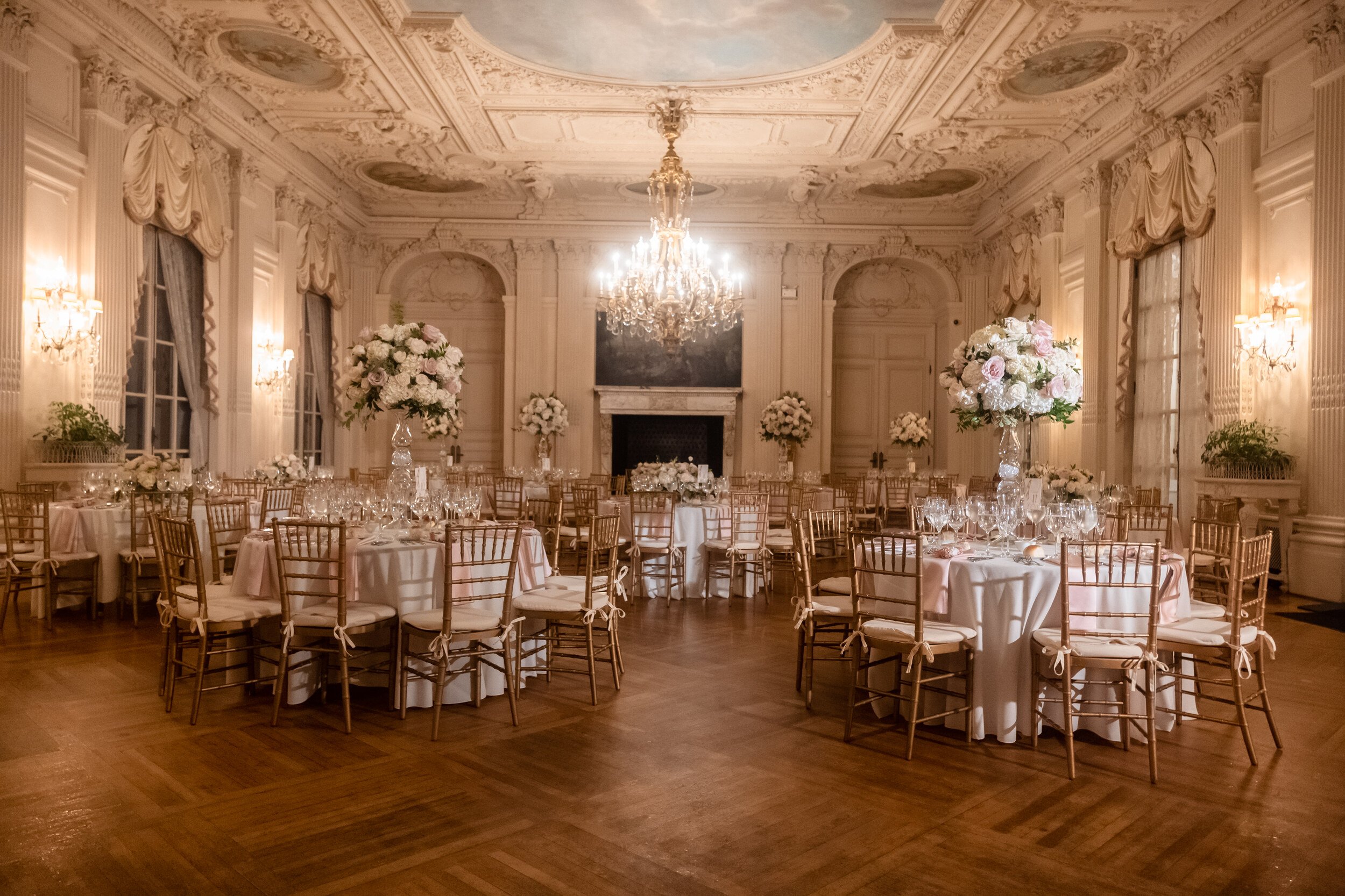 Empty ballroom set for a wedding reception at Rosecliff: a chandelier and painted ceiling above round tables in white linen with gold chiavari chairs and blush floral arrangements.