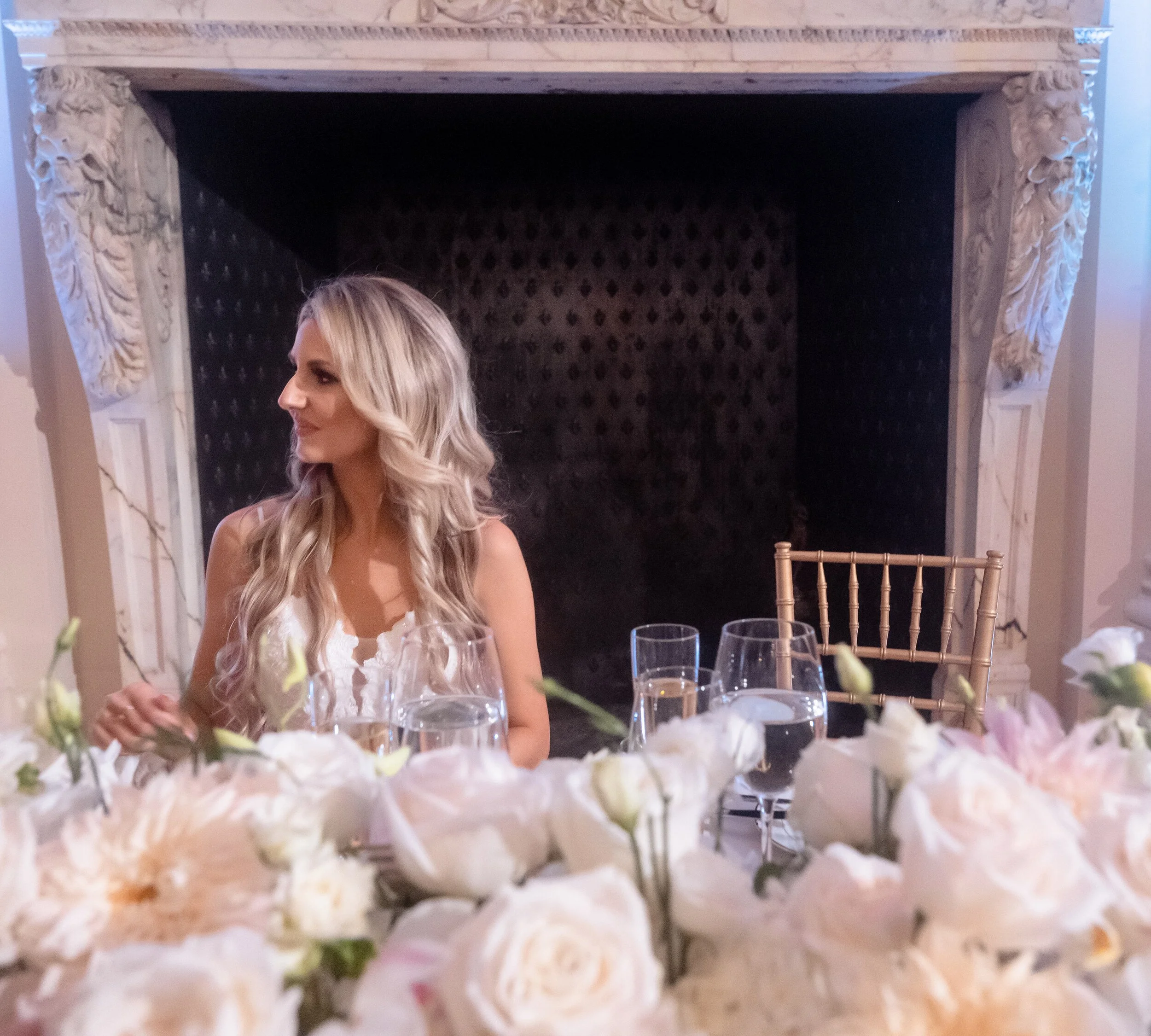 Bride alone in profile at a table set with blush and cream centerpieces, an ornate carved marble alcove behind her, Rosecliff.