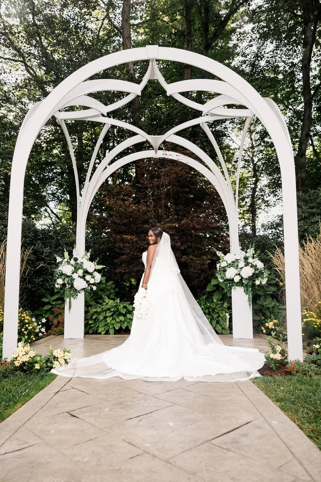 Avenir wedding videographer, Walpole, MA — bride framed beneath the white wrought arbor on the stone ceremony aisle