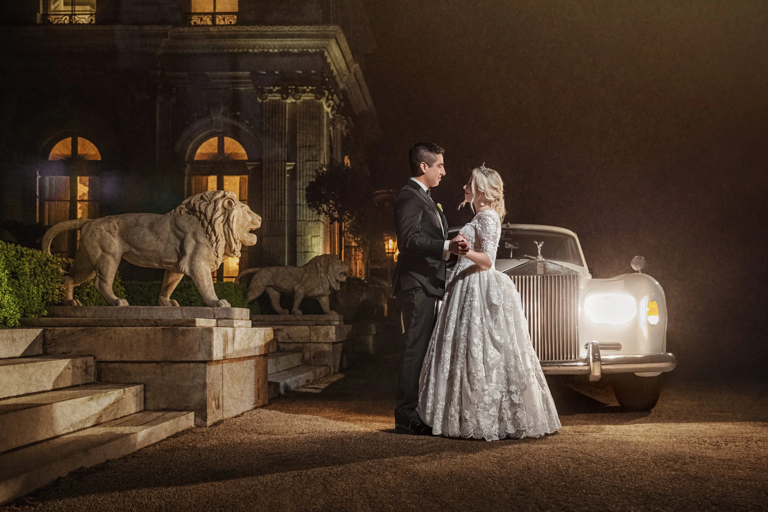 A couple beside a vintage Rolls-Royce at the entrance to Rosecliff Mansion after dark, the stone lions and the lit windows of the house behind them, Newport, Rhode Island