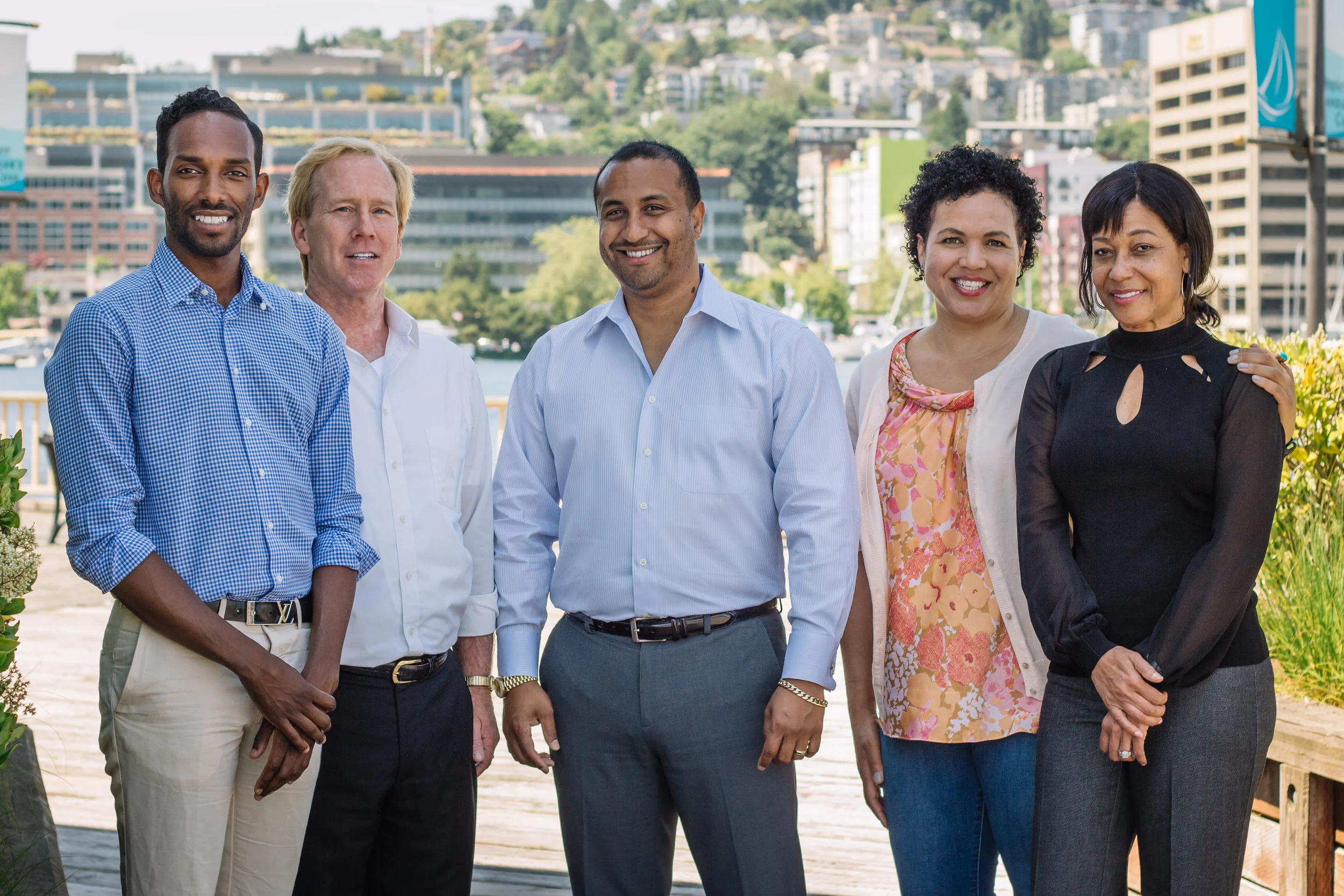 A diverse group of five adults standing outdoors by a waterfront with buildings and hills in the background, smiling at the camera.
