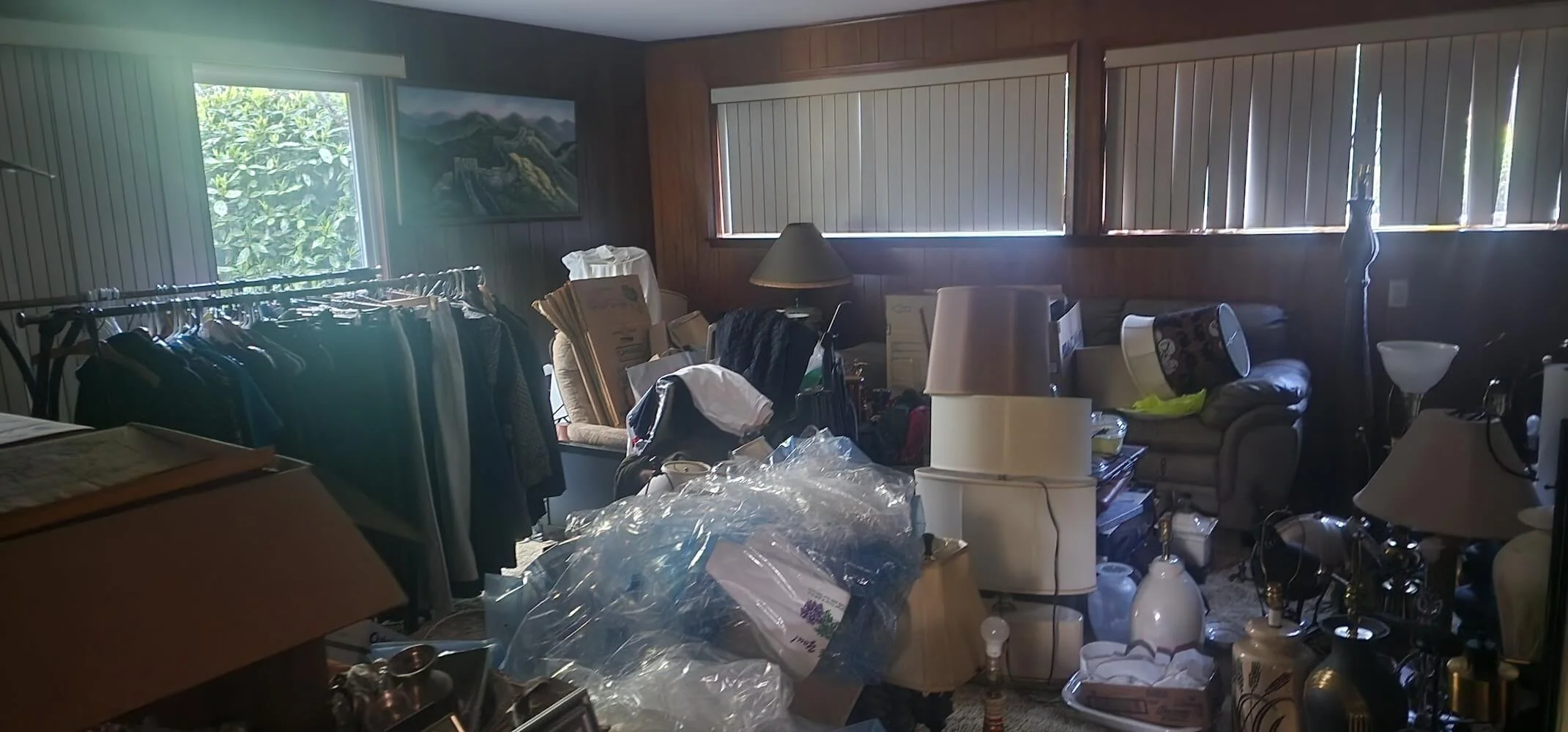 Room filled with clutter including clothes on a rack, boxes, lamps, furniture, and various household items.