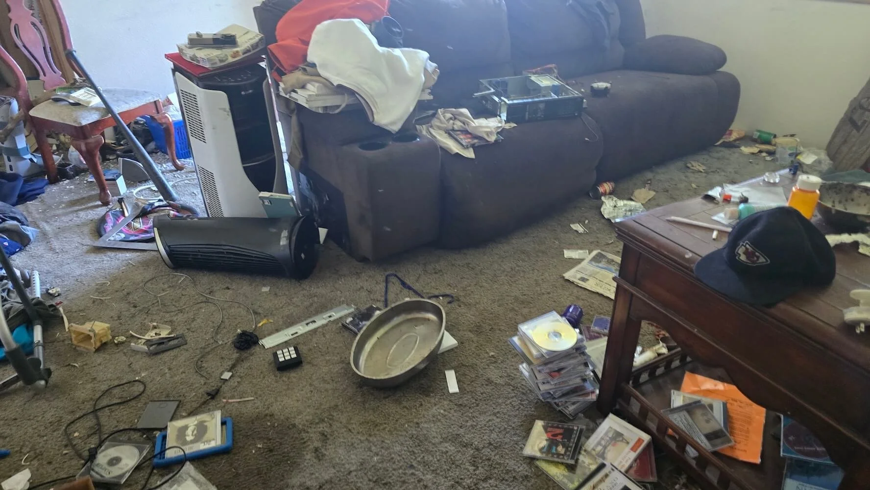 Living room in disarray with scattered clutter including a fallen tabletop, CDs, DVDs, trash, dirty laundry, and broken or misplaced items.