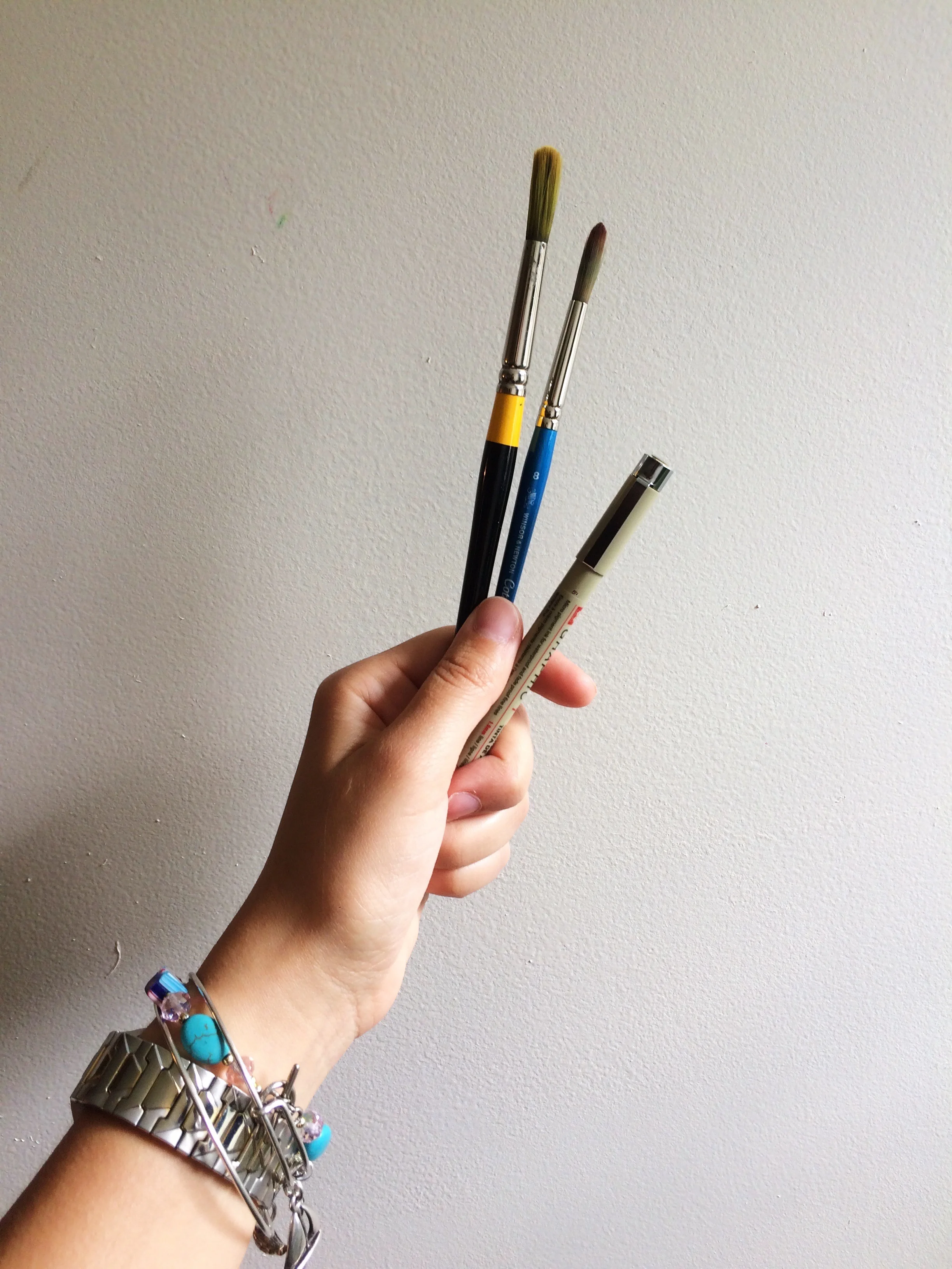 A hand holding three paintbrushes against a white wall, with a bracelet on the wrist.