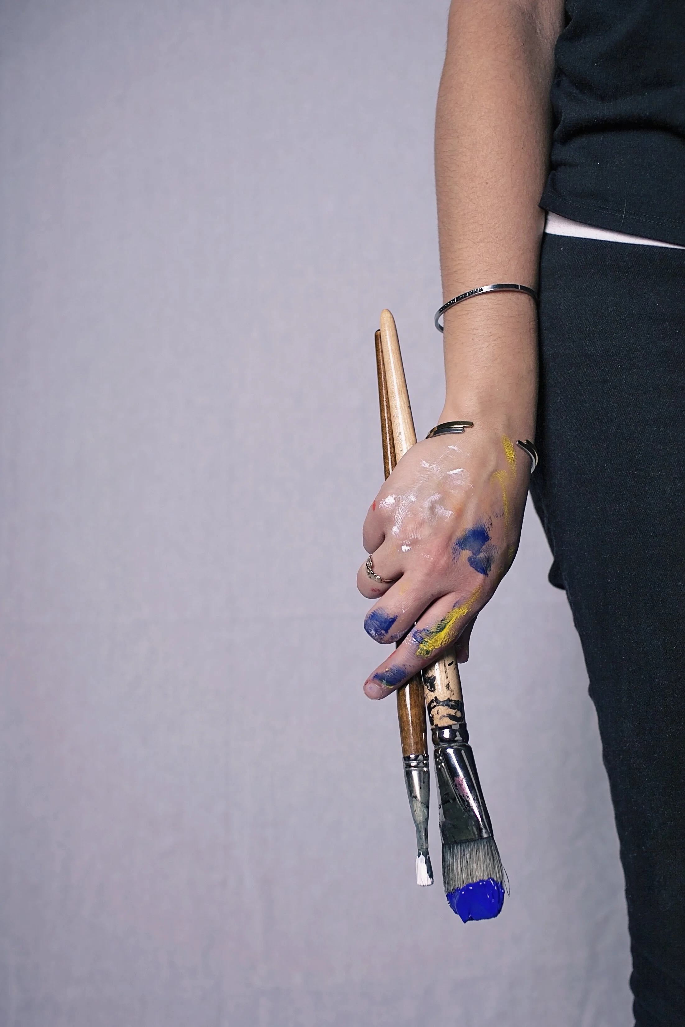 Close-up of a person's hand holding three paintbrushes, with their arm and side of their body visible. The hand and arm are covered in splashes of colorful paint, and the person is wearing rings and a bracelet, with a gray background.