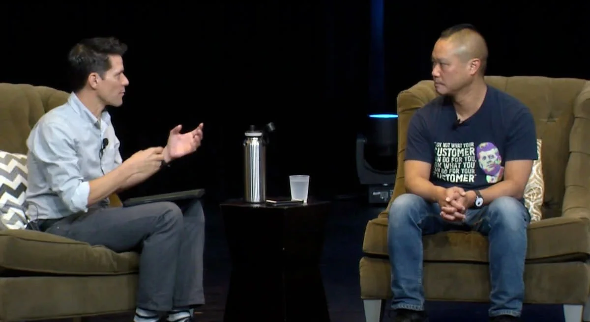 Two men sitting on brown couches engaging in conversation during an interview on a dark stage. One man wears a light-colored shirt, and the other wears a dark T-shirt and jeans. A small black table with a water bottle, cup, and an electronic device is between them.