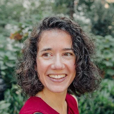 A smiling woman with curly dark hair wearing a red top outdoors with greenery in the background.