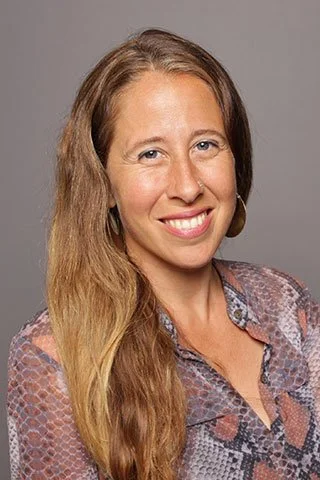 A woman with long, wavy brown hair smiling at the camera, wearing a patterned blouse and hoop earrings against a gray background.