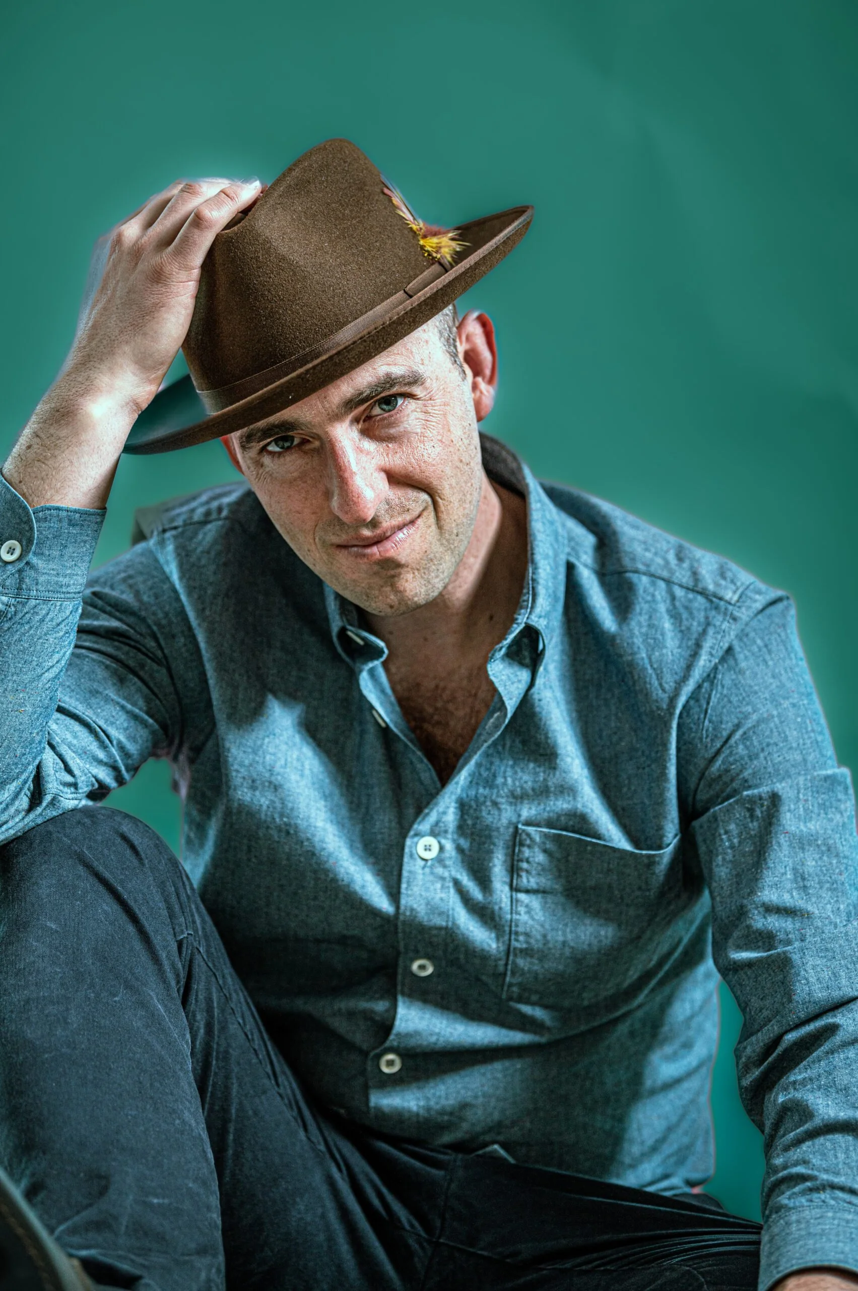 A man wearing a denim shirt is sitting and tilting his head, holding a brown hat with a feather on top.