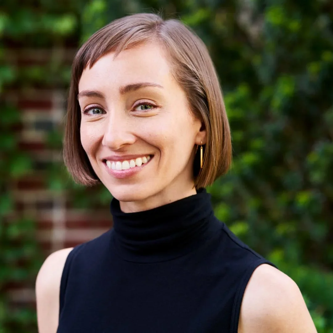 A woman with short brown hair, smiling, wearing a black sleeveless turtleneck top, outdoors with green foliage in the background.