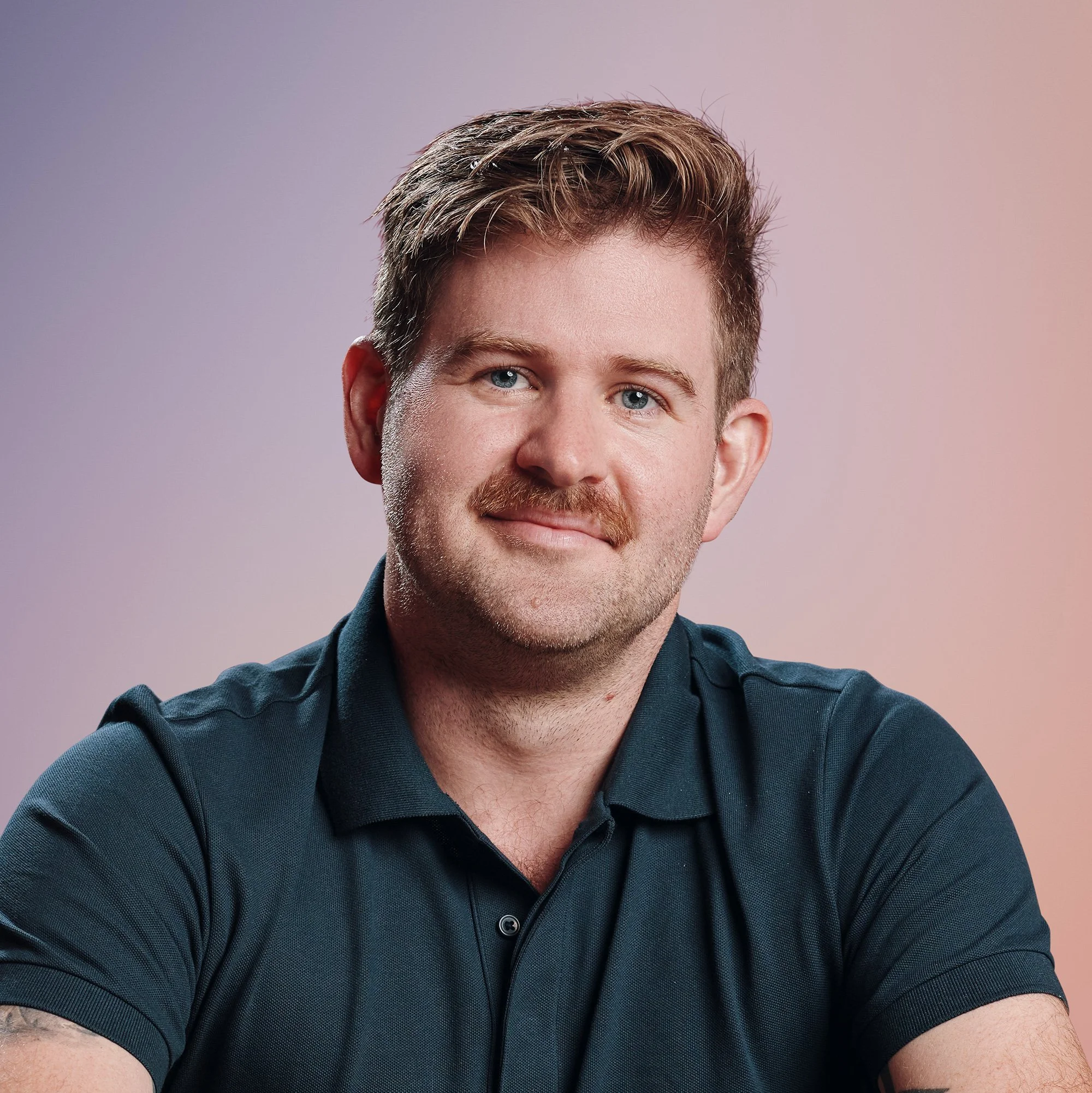 Portrait of a man with light skin, short brown hair, a mustache, and a light beard, wearing a dark blue polo shirt, smiling with a pastel pink and purple gradient background.