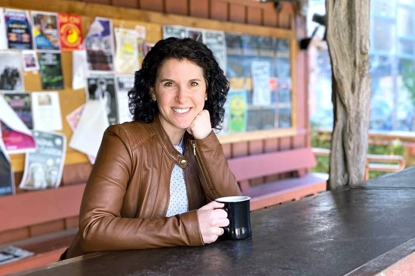 A woman with curly dark hair, smiling, sitting at an outdoor table with a coffee mug, in front of a bulletin board with notices, in a wooden structure.