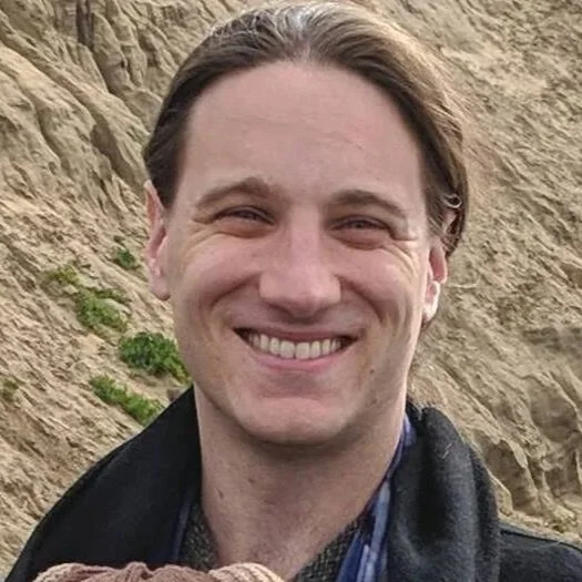 A smiling man with short brown hair stands outdoors in front of a rocky, sandy hillside with sparse green vegetation.