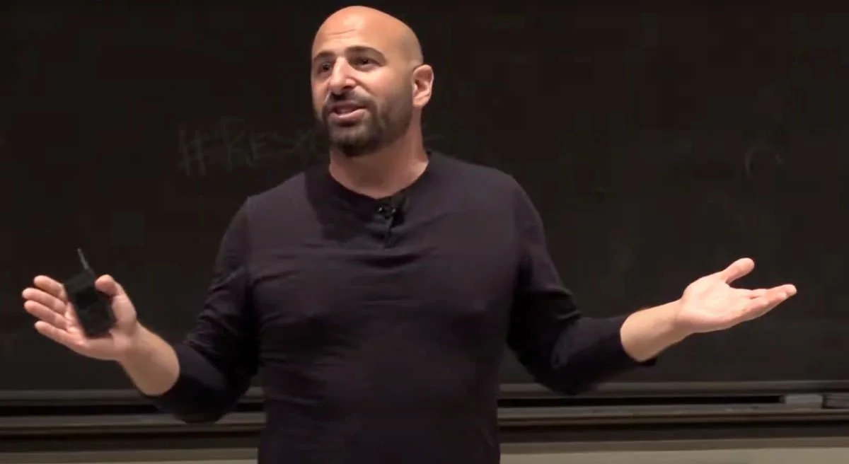 A man with a beard and no hair, wearing a black long-sleeve shirt, stands in front of a blackboard or dark background, gesturing with both hands while speaking.