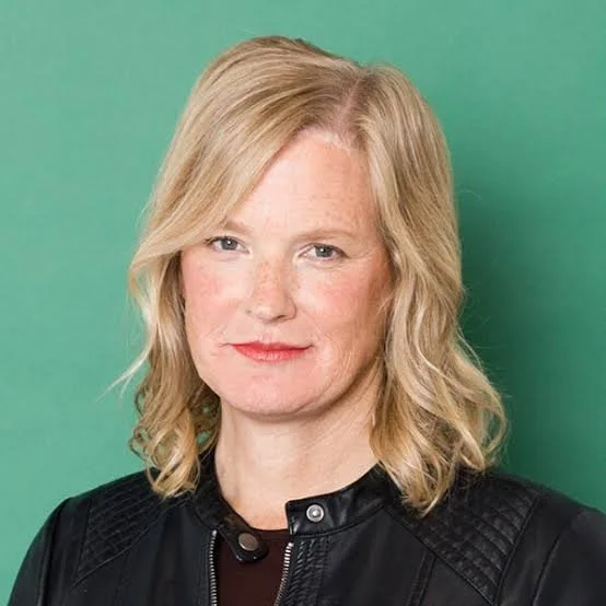 A woman with blonde, wavy hair and fair skin, wearing a black leather jacket, stands against a green background.